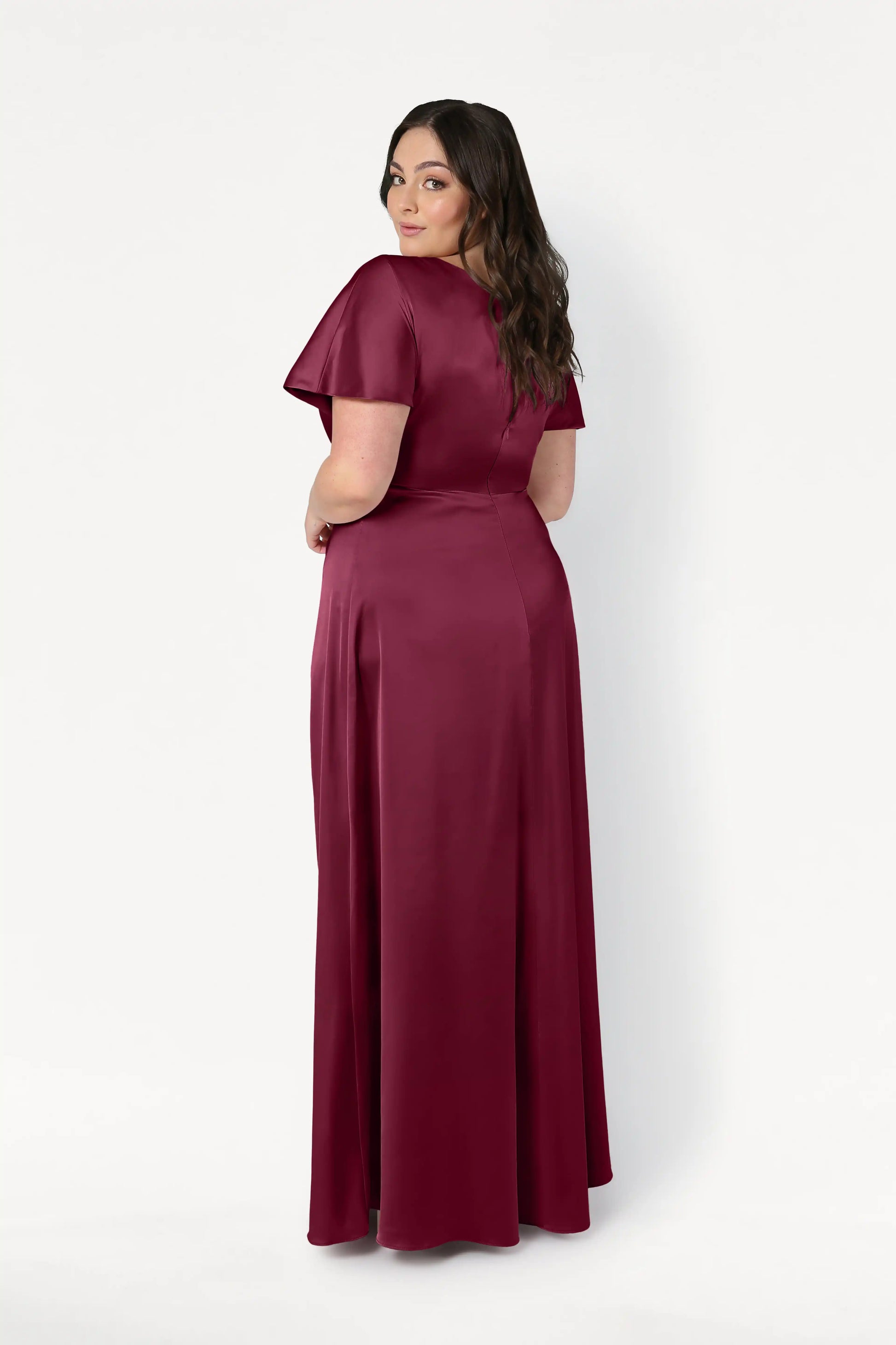A woman with long dark hair looks over her shoulder, wearing the TH&TH Camilla Bridesmaid Dress in Merlot Satin—a modern, floor-length, short-sleeved gown—posing against a plain white background.