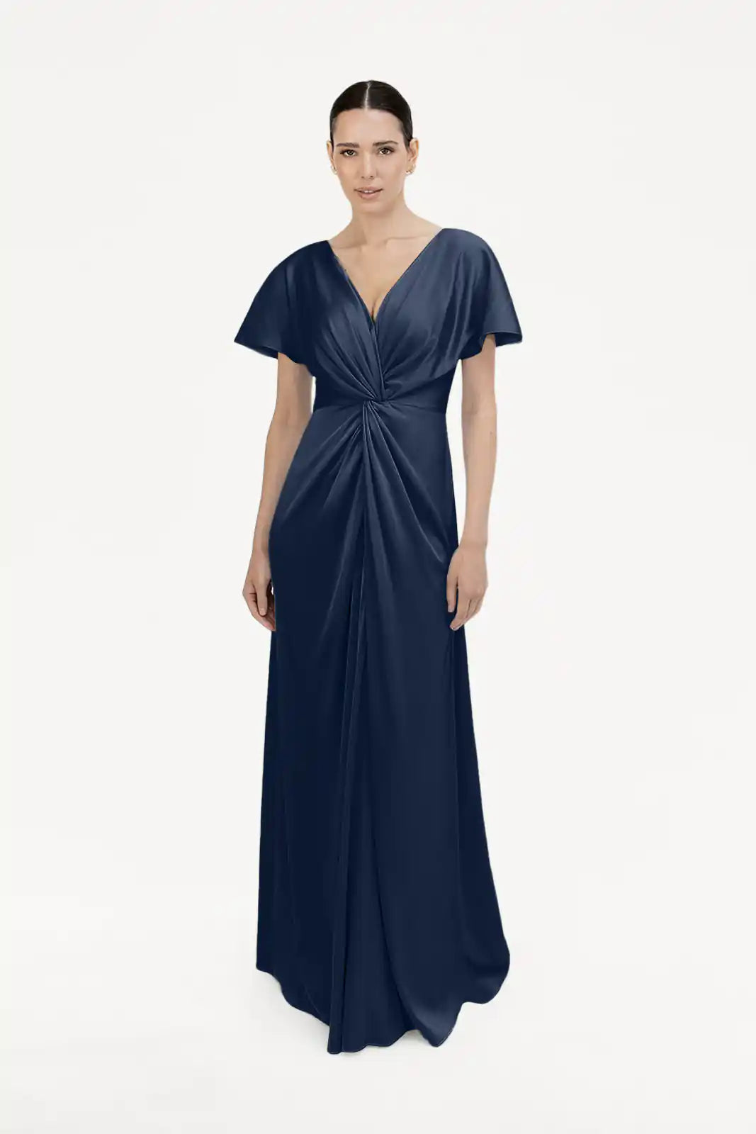 A woman stands against a plain white background wearing the TH&TH Camilla Bridesmaid Dress in Navy Satin, a size-inclusive, floor-length gown featuring short sleeves, a deep V-neckline, and a twist-knot bodice at the waist.