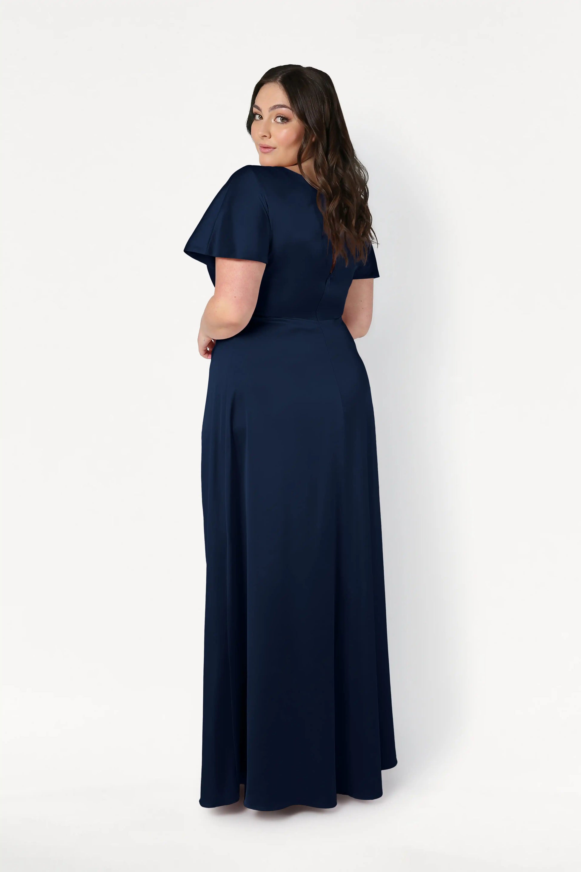 A woman with long wavy brown hair wears the size-inclusive Camilla Bridesmaid Dress in Navy Satin by TH&TH, a short-sleeve, floor-length gown, as she stands facing away and looks over her shoulder against a plain white background.