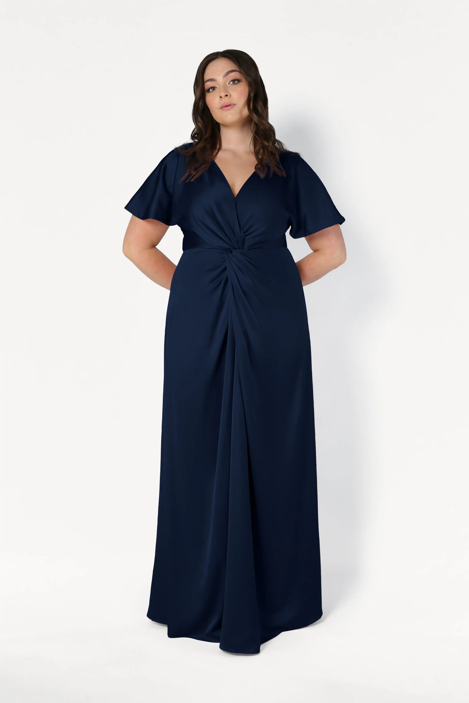 A woman poses against a white background wearing the TH&TH Camilla Bridesmaid Dress in Navy Satin—a size-inclusive, floor-length gown with short sleeves, a twist-knot waist, and a V-neckline. Her hands are behind her back.