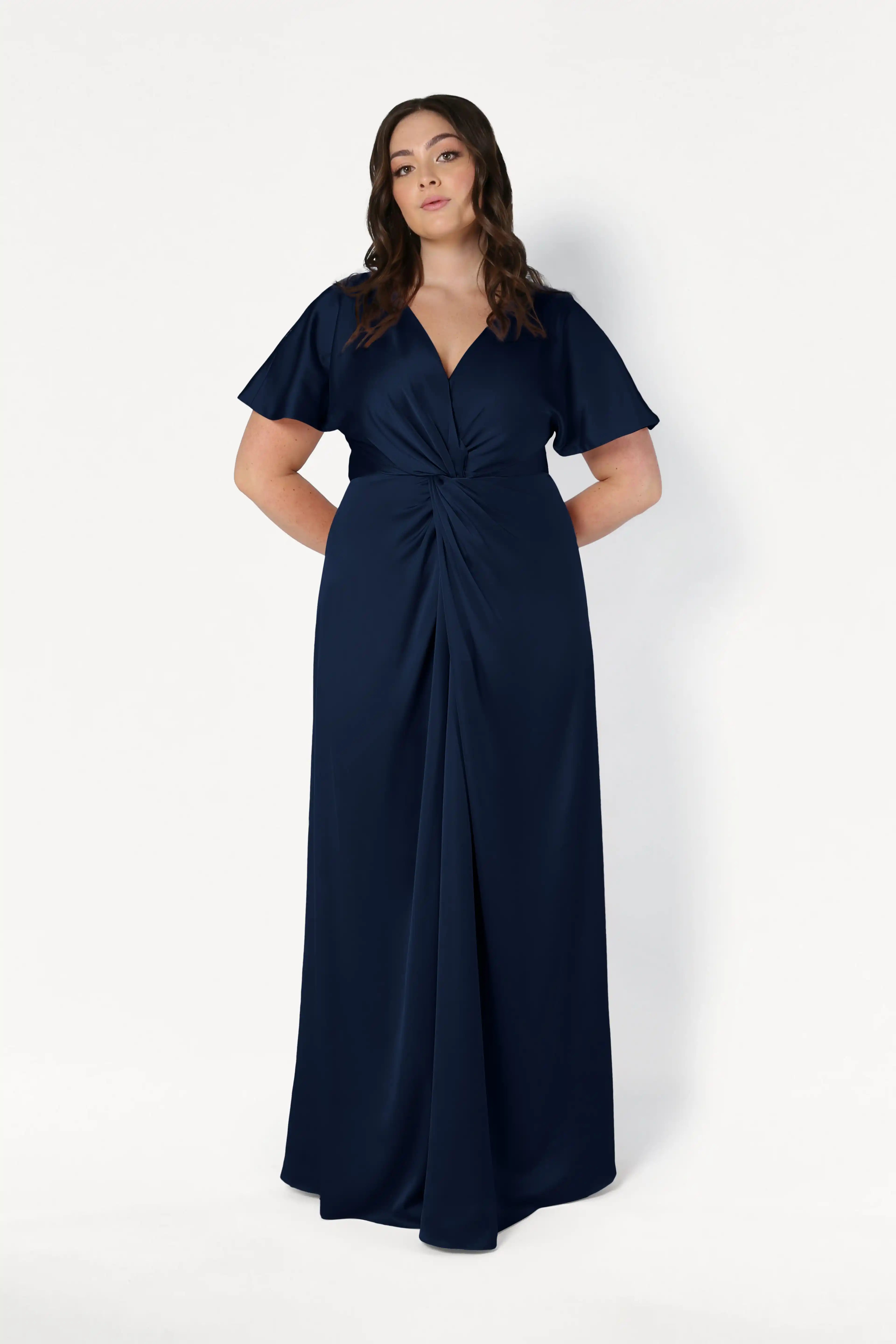 A woman poses against a white background wearing the TH&TH Camilla Bridesmaid Dress in Navy Satin—a size-inclusive, floor-length gown with short sleeves, a twist-knot waist, and a V-neckline. Her hands are behind her back.