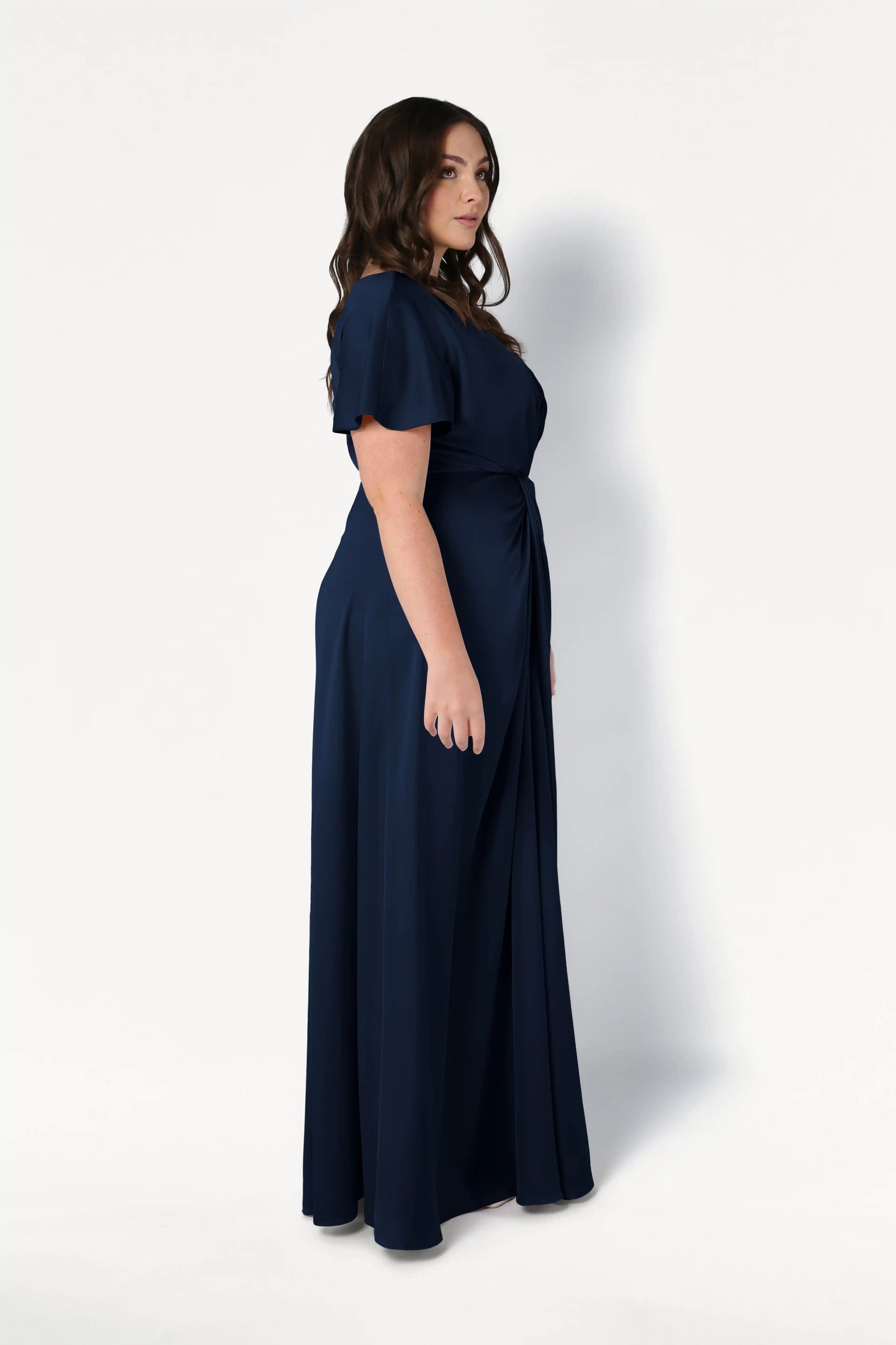 A woman with wavy brown hair stands in profile, modeling the TH&TH Camilla Bridesmaid Dress in Navy Satin—a size-inclusive, floor-length gown with short sleeves and a twist-knot bodice—against a plain white background.