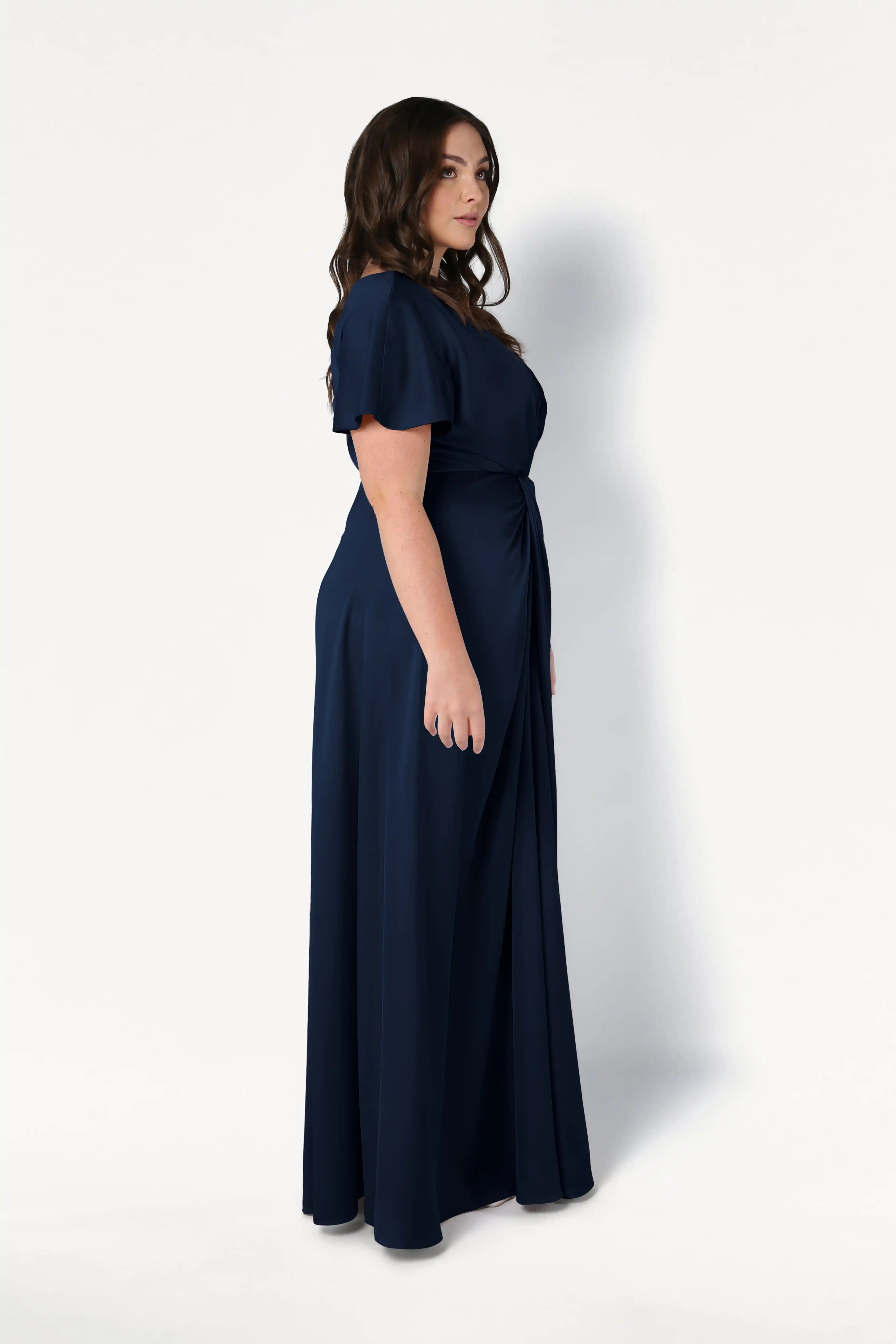 A woman with wavy brown hair stands in profile, modeling the TH&TH Camilla Bridesmaid Dress in Navy Satin—a size-inclusive, floor-length gown with short sleeves and a twist-knot bodice—against a plain white background.