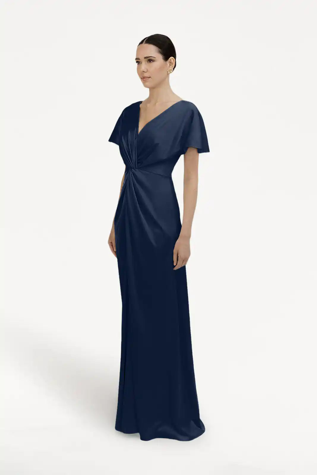 A woman stands against a white background wearing the Camilla Bridesmaid Dress in Navy Satin by TH&TH, a size-inclusive gown with short sleeves, V-neckline, and gathered twist-knot bodice. Her hair is styled in a neat bun.