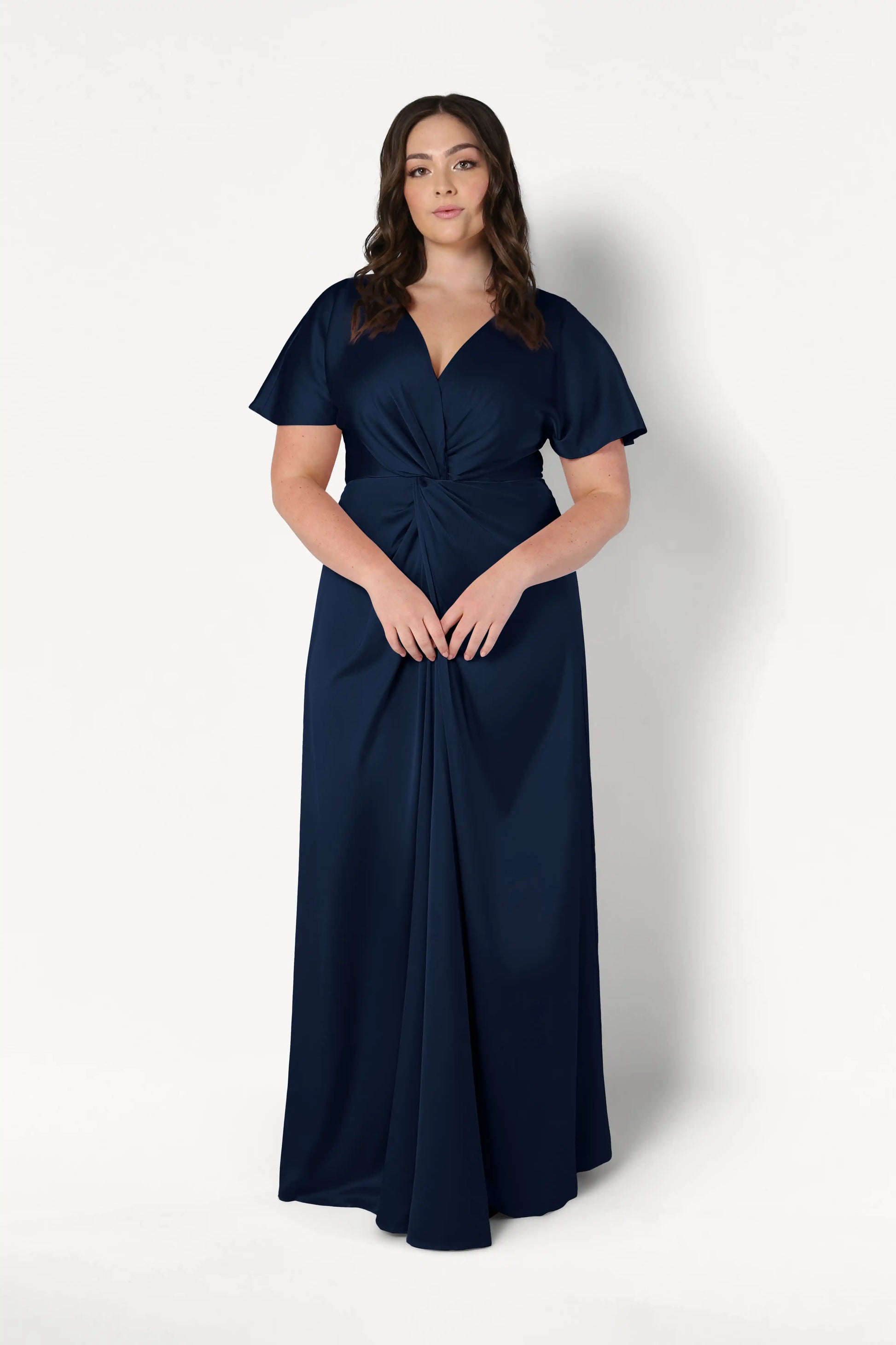 A woman wears the Camilla Bridesmaid Dress in Navy Satin by TH&TH—a size-inclusive, floor-length gown with short flutter sleeves and a twist-knot bodice—posing against a plain white background.
