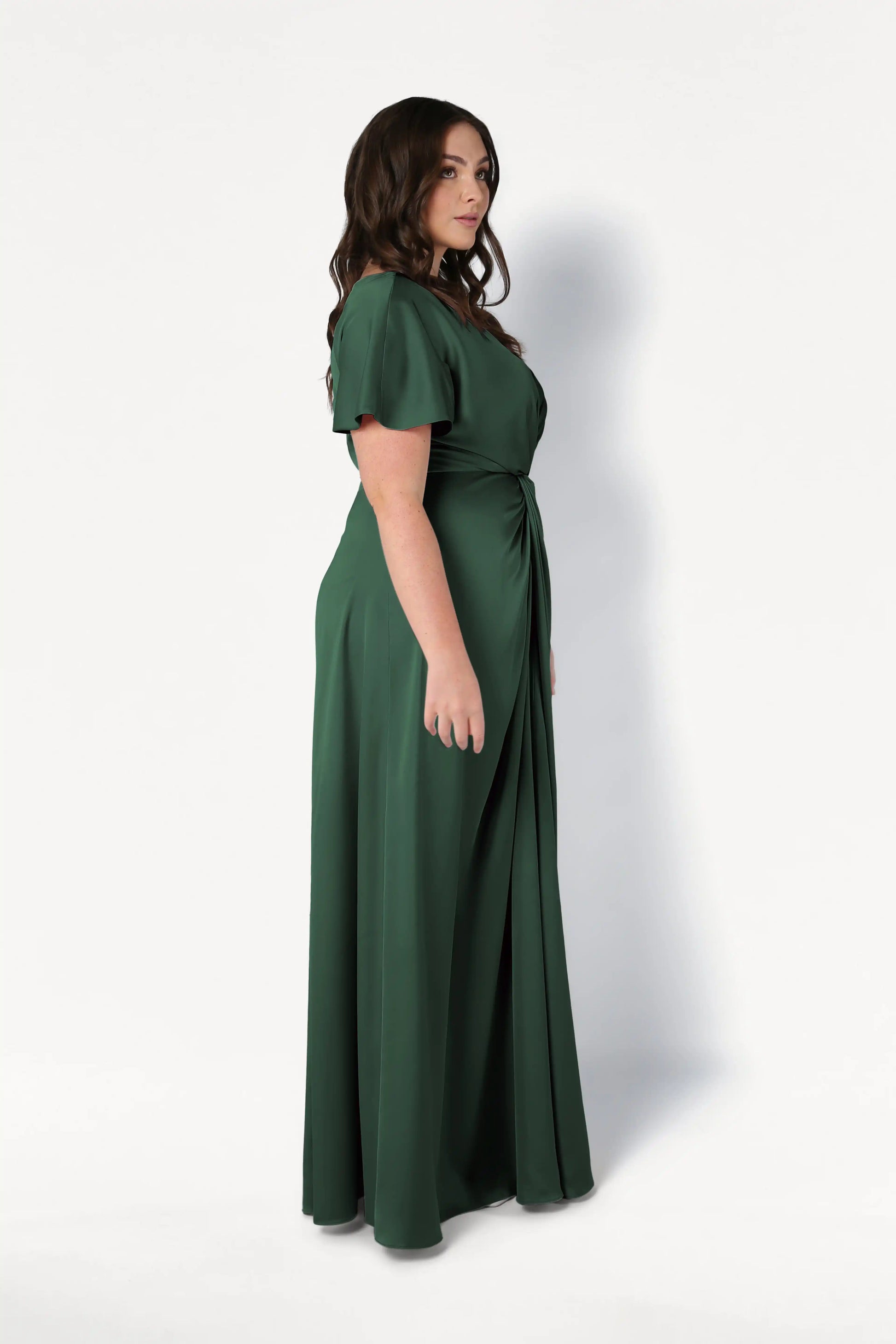 A woman with wavy brown hair stands in profile, modeling the TH&TH Camilla Bridesmaid Dress in Olive Grove Satin—a modern, floor-length gown with short sleeves and a gathered waist—against a plain white background.