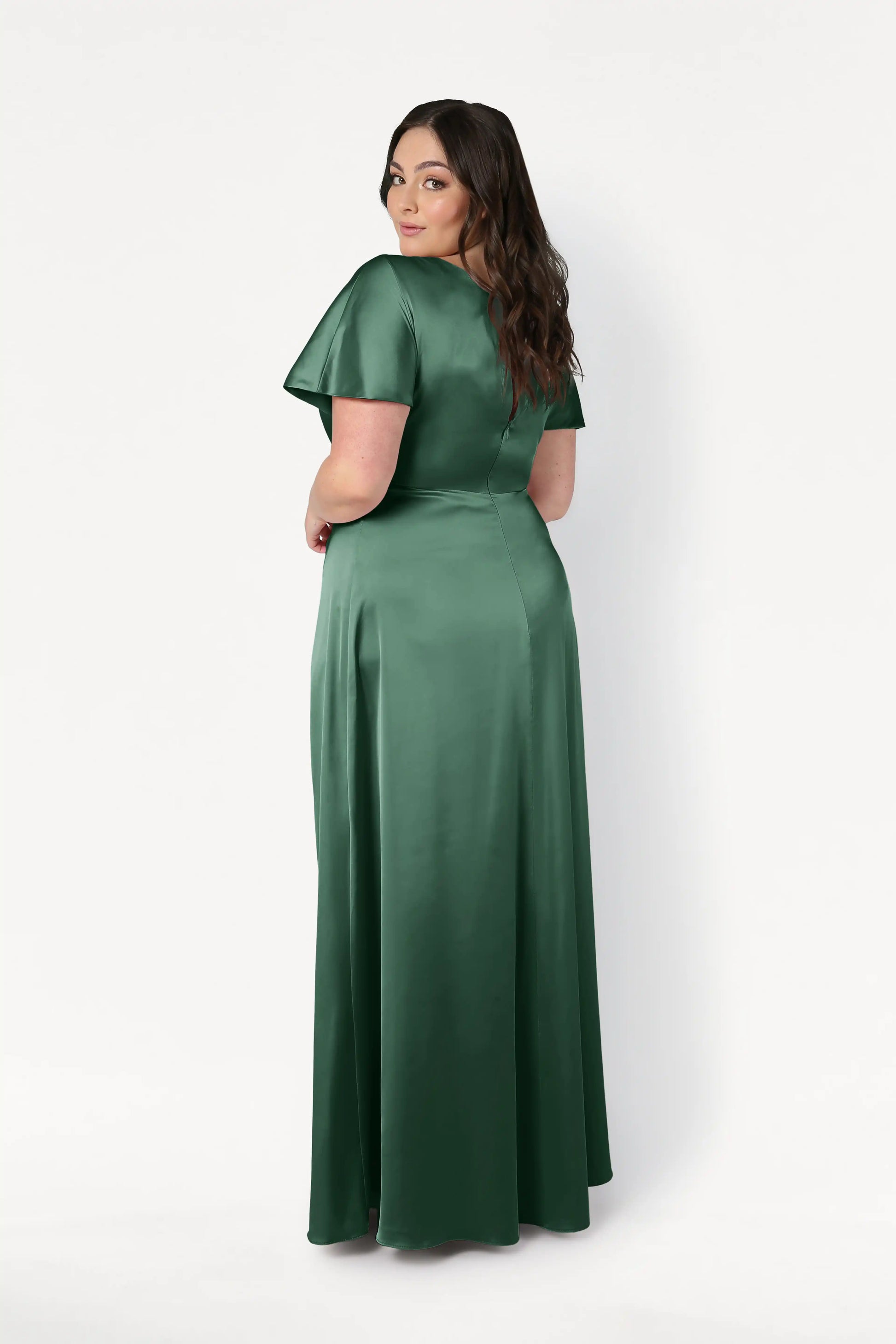 A woman with long brown hair wears the TH&TH Camilla Bridesmaid Dress in Olive Grove Satin, a floor-length, short-sleeve gown. She stands against a plain white background, facing away and looking back over her shoulder.