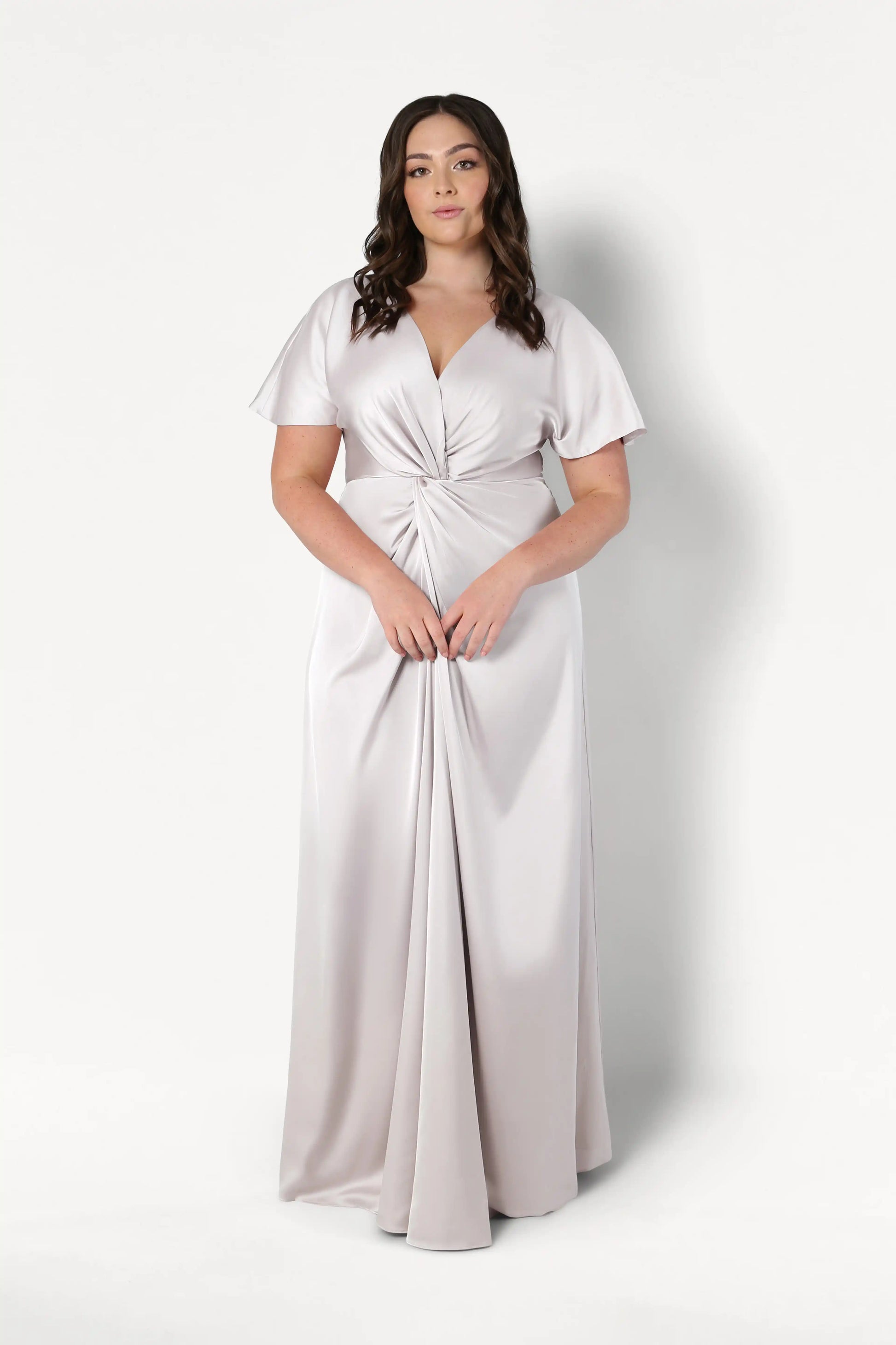 A woman with long brown hair wears the TH&TH Camilla Bridesmaid Dress in Oyster Satin, featuring short flutter sleeves and a twist waist detail, standing against a plain white background.