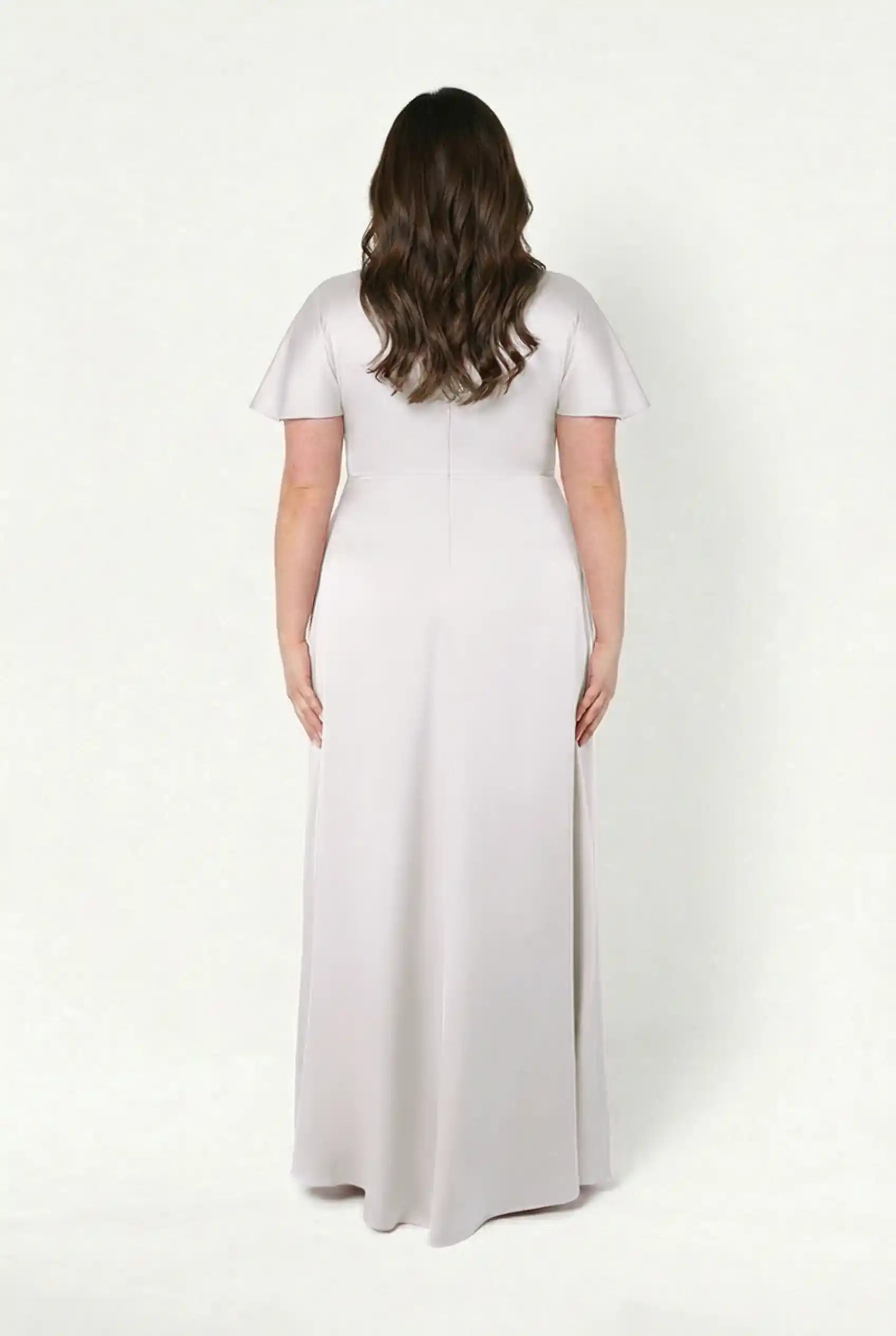 A woman with long wavy brown hair stands with her back to the camera, wearing the TH&TH Camilla Bridesmaid Dress in Oyster Satin, a floor-length, short-sleeve gown, against a plain white background.
