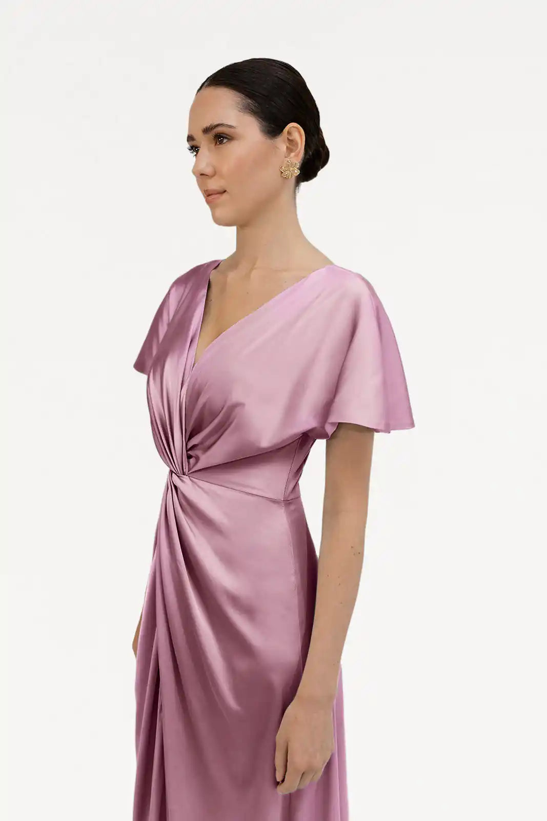 A woman with dark hair in a low bun models the Camilla Bridesmaid Dress in Rose Pink Satin by TH&TH, featuring short flutter sleeves and gathered waist detailing. She stands against a plain white background, looking to the left.