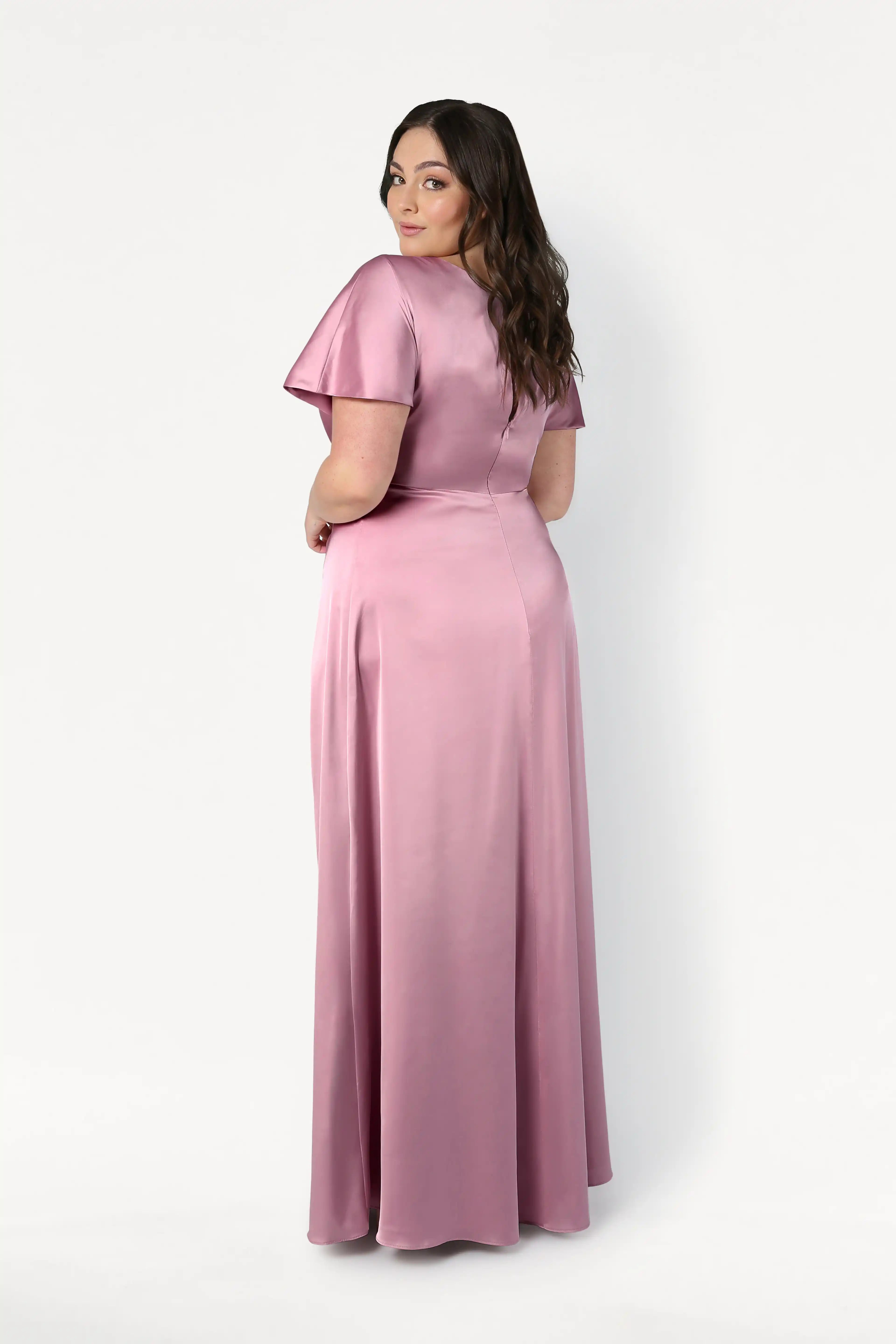 A woman with long brown hair models the TH&TH Camilla Bridesmaid Dress in Rose Pink Satin, a floor-length gown with short sleeves and a twist-knot bodice. She stands turned to the side, looking over her shoulder against a white background.