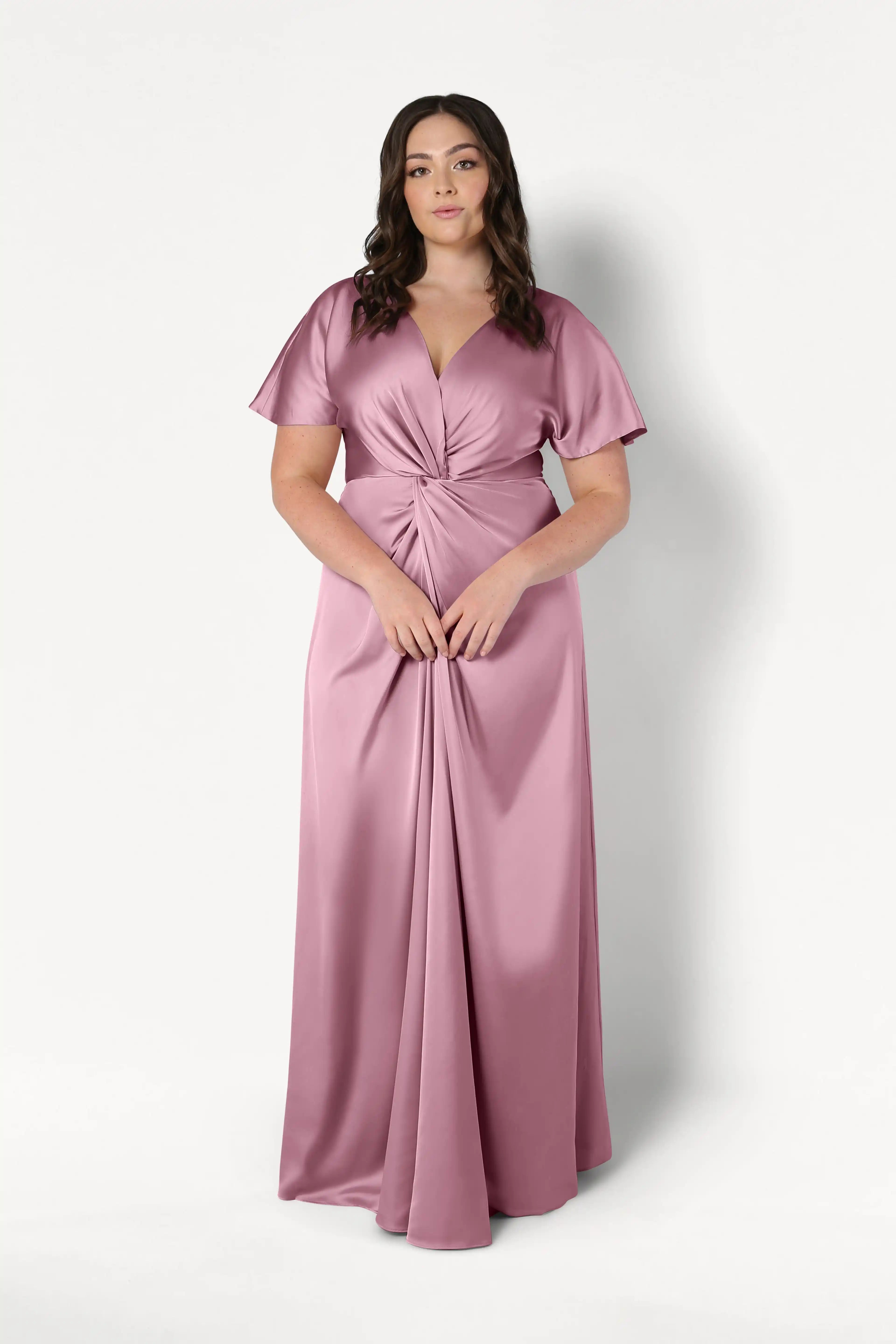 A woman poses against a white backdrop in the TH&TH Camilla Bridesmaid Dress in rose pink satin, featuring a floor-length silhouette, short sleeves, twist-knot bodice, V-neckline, flattering waist detail, and inclusive sizing.