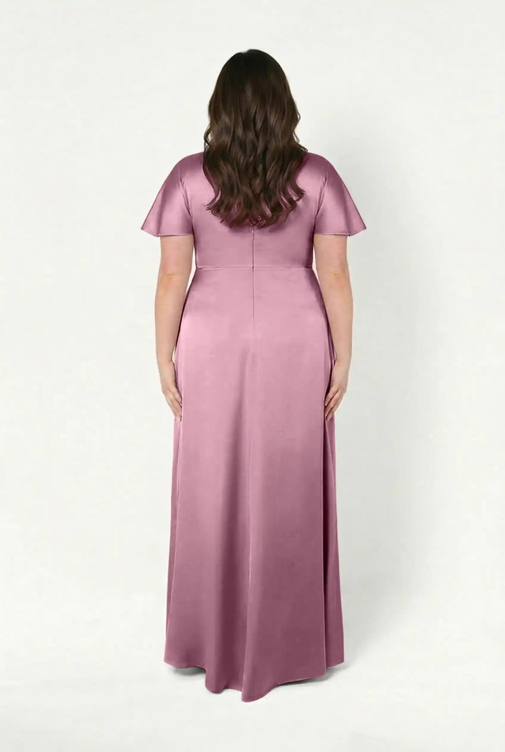 A woman with long brown hair stands facing away in the TH&TH Camilla Bridesmaid Dress in rose pink satin, featuring a twist-knot bodice and short sleeves. This elegant, floor-length style is size-inclusive, shown against a plain white background.