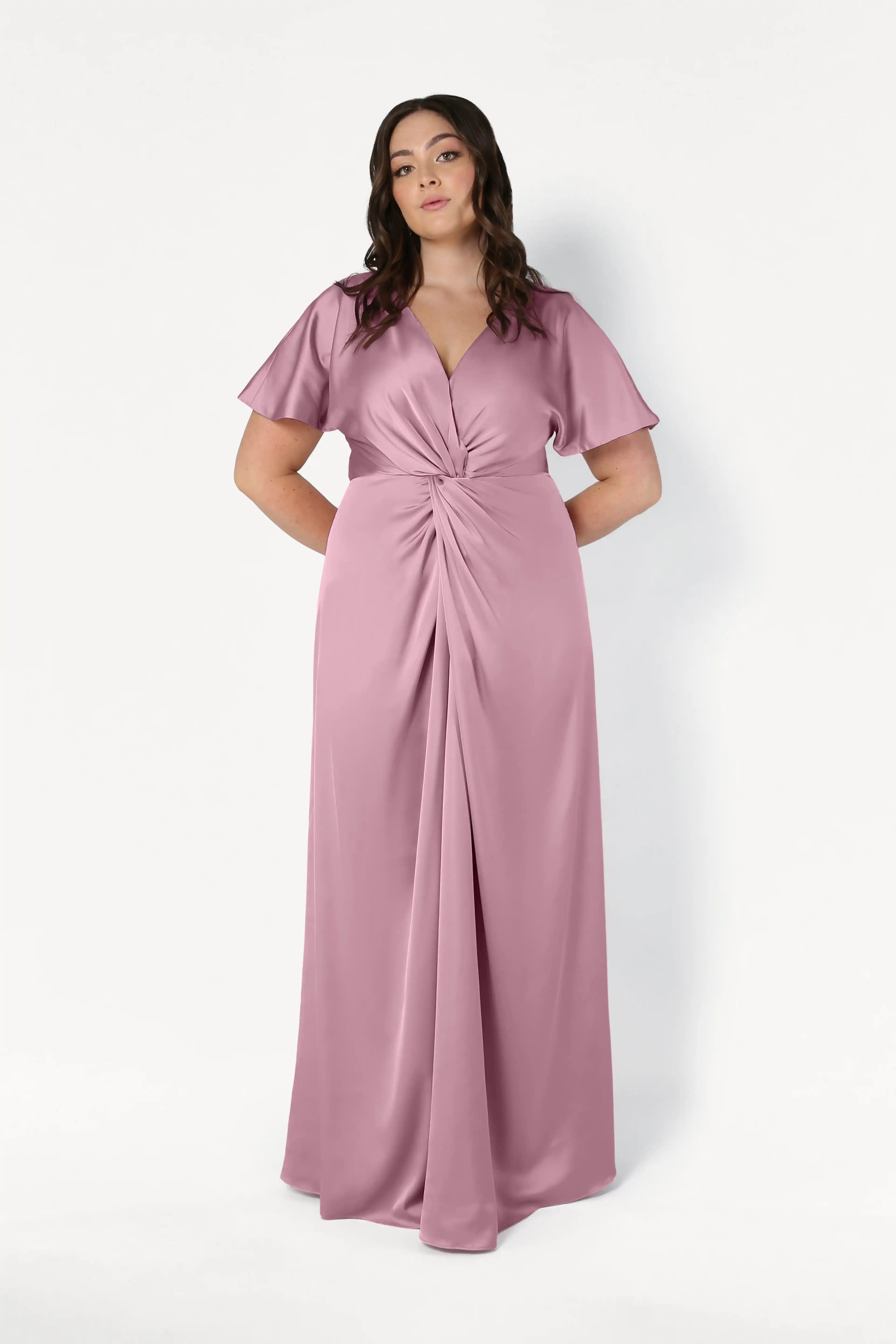 A woman wears the TH&TH Camilla Bridesmaid Dress in rose pink satin—a floor-length, size-inclusive gown with short flutter sleeves and a twist-knot bodice—standing confidently against a white background, hands behind her back.