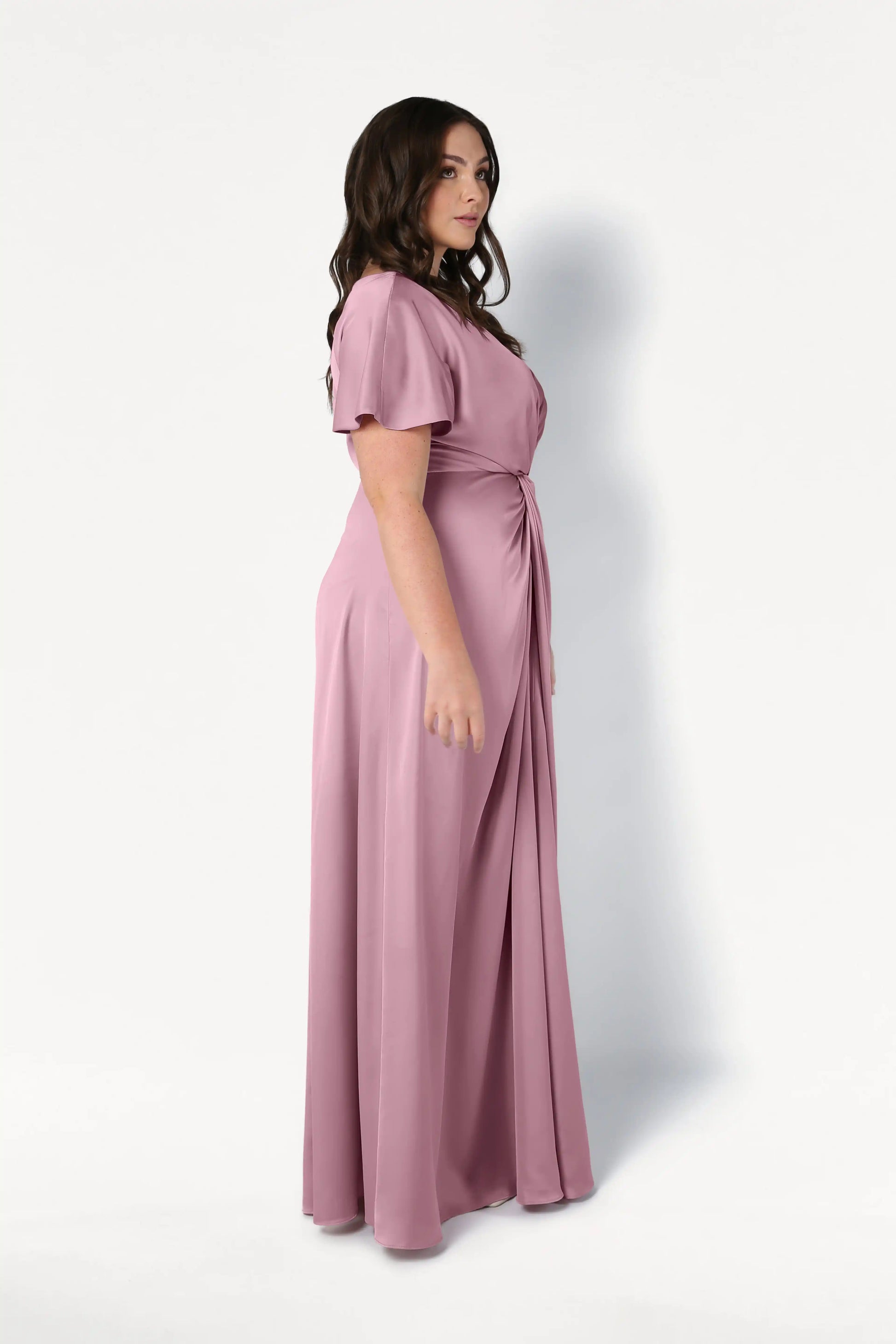 A woman with long dark hair stands in profile against a white background, wearing the Camilla Bridesmaid Dress in Rose Pink Satin by TH&TH, a floor-length, size-inclusive gown with a gathered waist and elegant short sleeves.