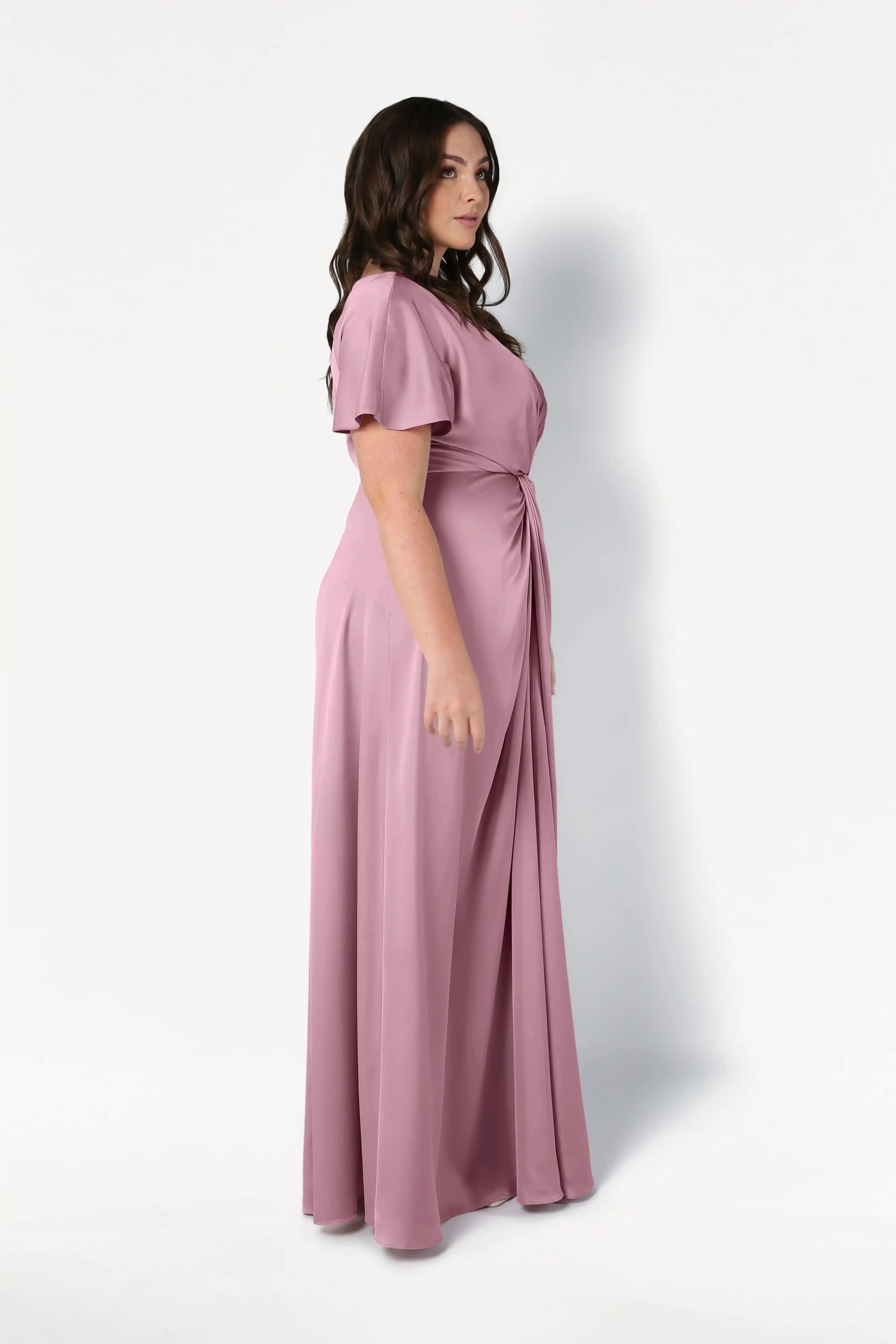 A woman with long dark hair stands in profile against a white background, wearing the Camilla Bridesmaid Dress in Rose Pink Satin by TH&TH, a floor-length, size-inclusive gown with a gathered waist and elegant short sleeves.