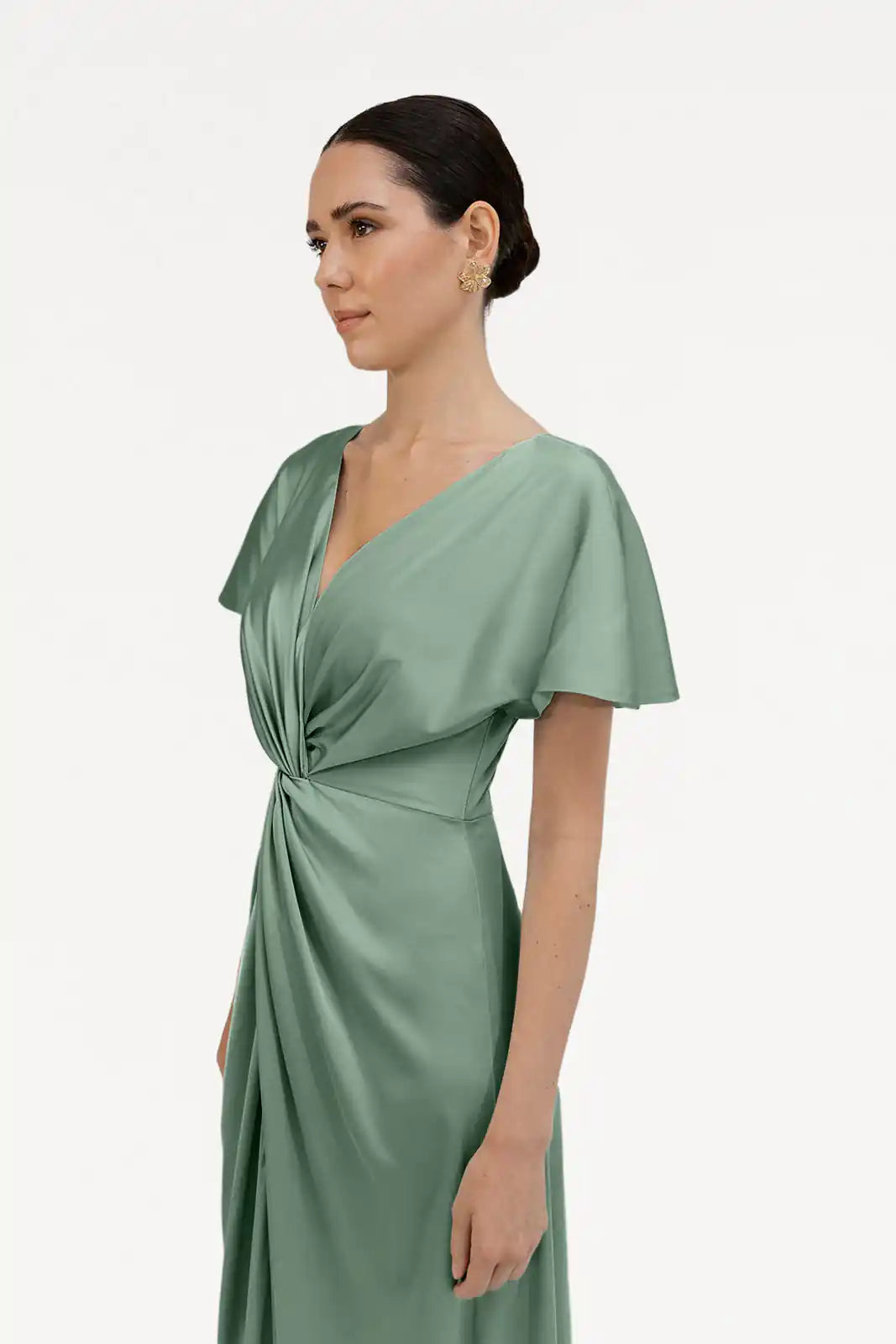 A woman with dark hair in a low bun wears the TH&TH Camilla Bridesmaid Dress in Sage Green Satin, featuring short sleeves and a twist-knot bodice, standing against a plain white background.