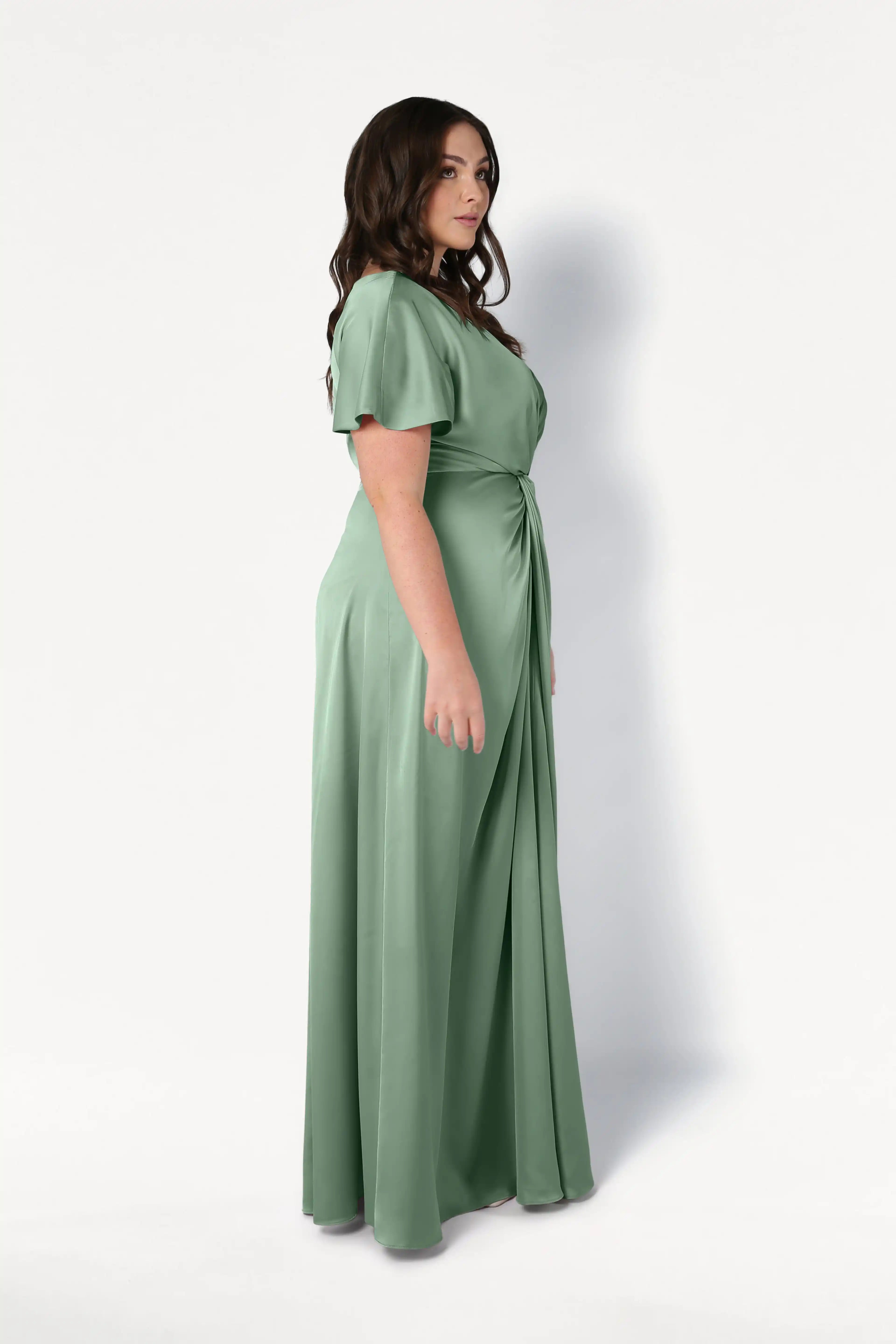 A woman with long dark hair stands in profile, wearing the TH&TH Camilla Bridesmaid Dress in sage green satin with short sleeves and a gathered waist, set against a plain white background.