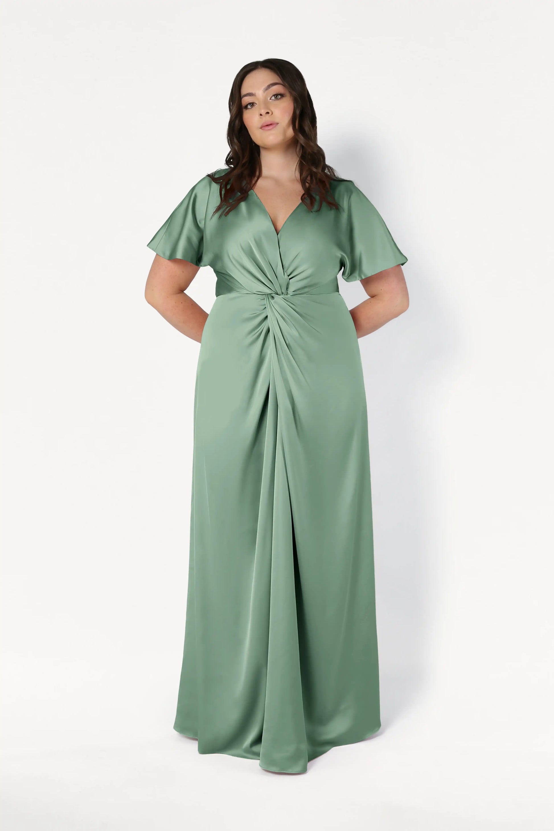 A woman with wavy brown hair stands against a plain white background, hands behind her back, wearing the TH&TH Camilla Bridesmaid Dress in size-inclusive sage green satin with flutter sleeves and a twist-knot bodice.