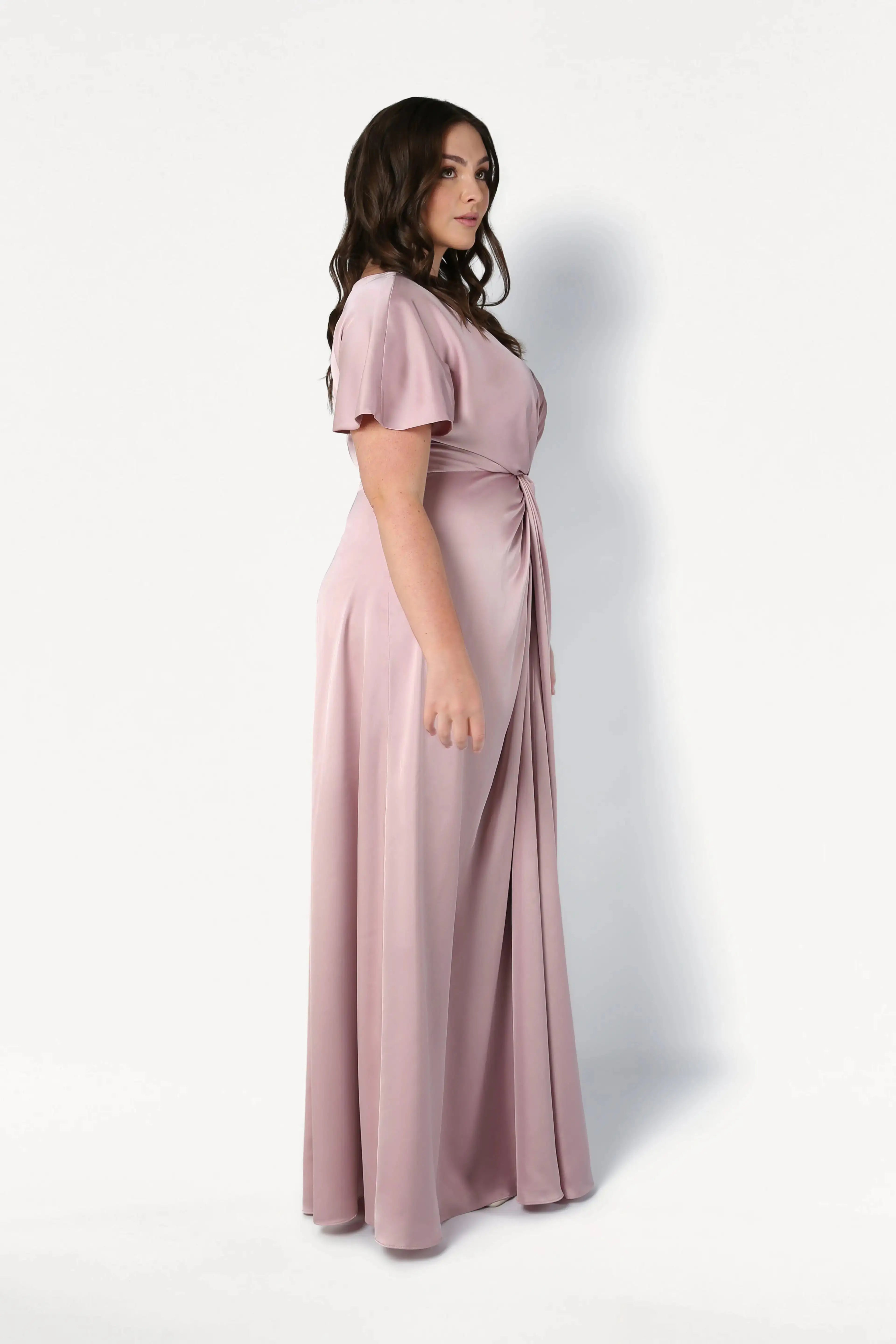 A woman with long dark hair stands sideways against a white background, wearing the TH&TH Camilla Bridesmaid Dress in smoked blush satin, a floor-length gown with short sleeves.