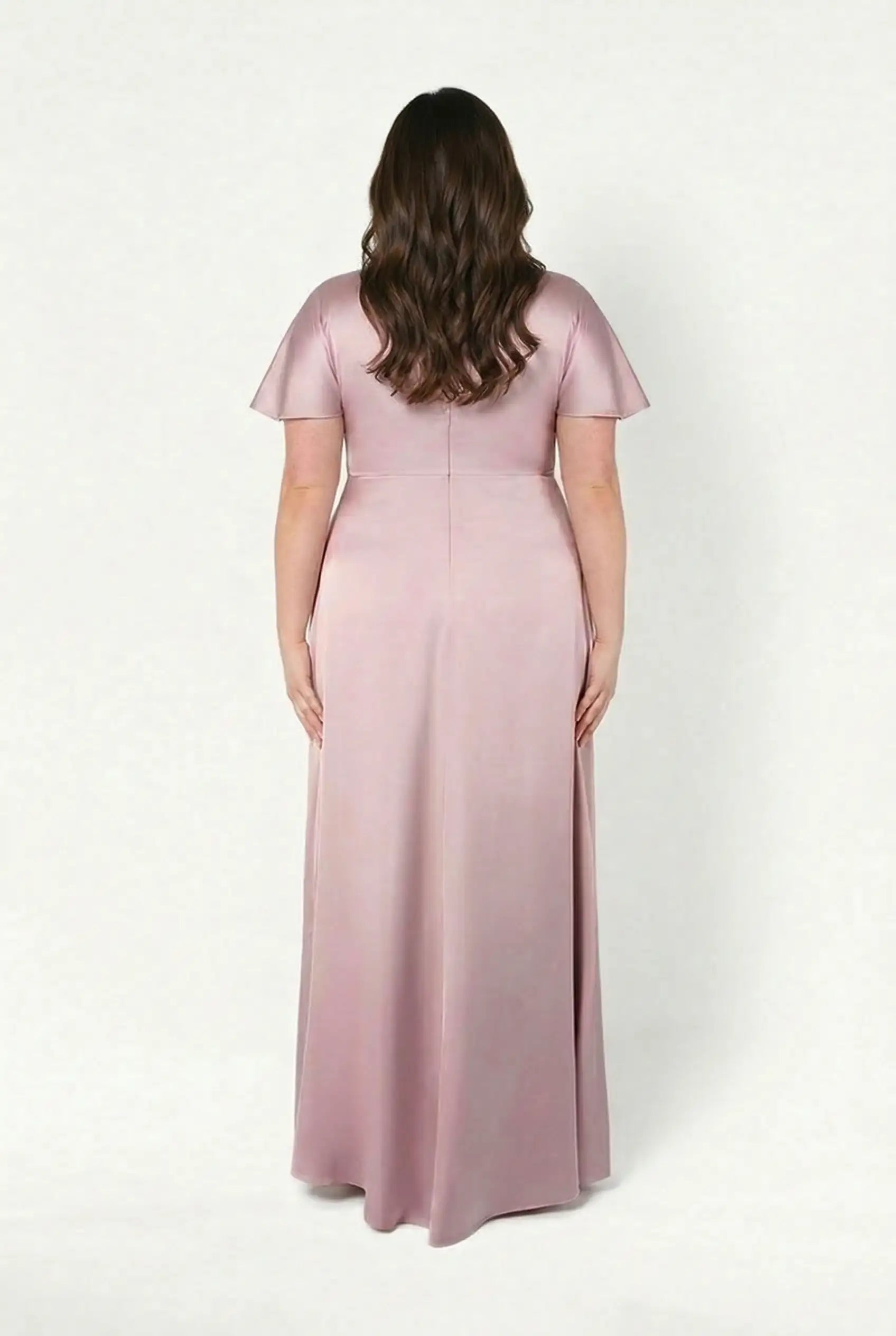 A woman with long, wavy brown hair faces away, wearing the TH&TH Camilla Bridesmaid Dress in Smoked Blush Satin—a modern, floor-length gown with short sleeves, perfect for any bridal party.