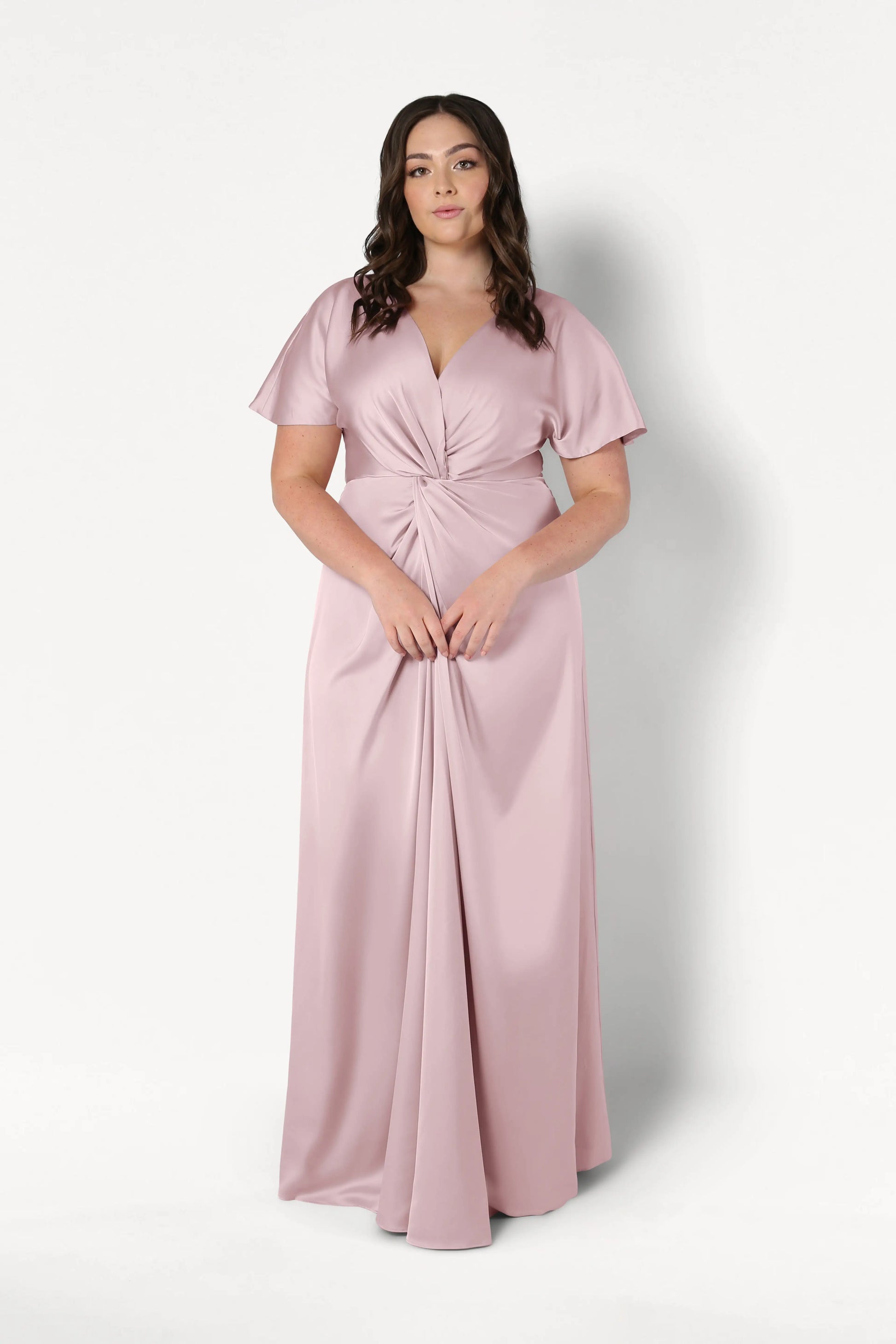 A woman with long brown hair wears the TH&TH Camilla Bridesmaid Dress in Smoked Blush Satin, a floor-length gown with a twisted knot waist detail, standing against a plain white background.