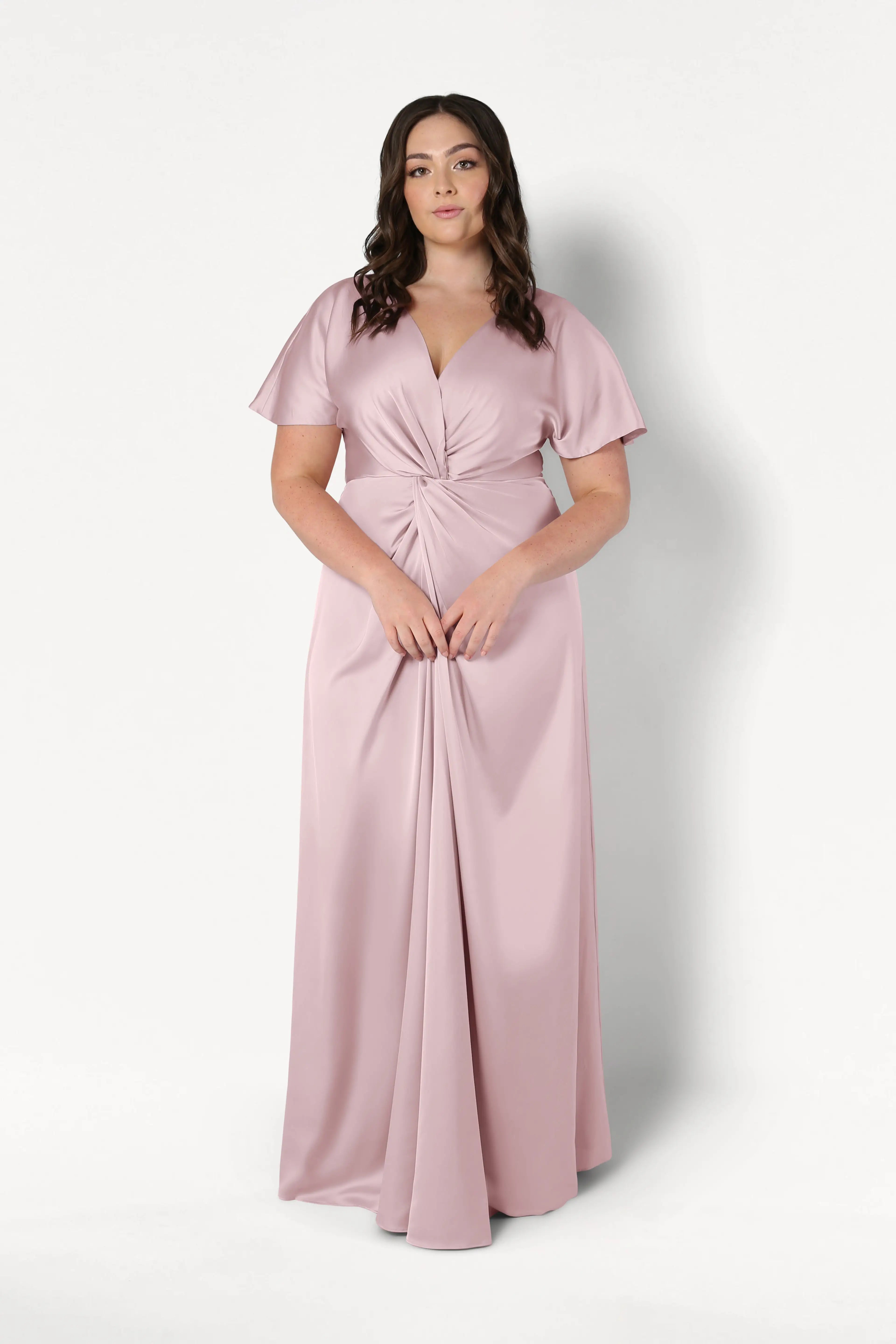 A woman with long brown hair wears the TH&TH Camilla Bridesmaid Dress in Smoked Blush Satin, a floor-length gown with a twisted knot waist detail, standing against a plain white background.