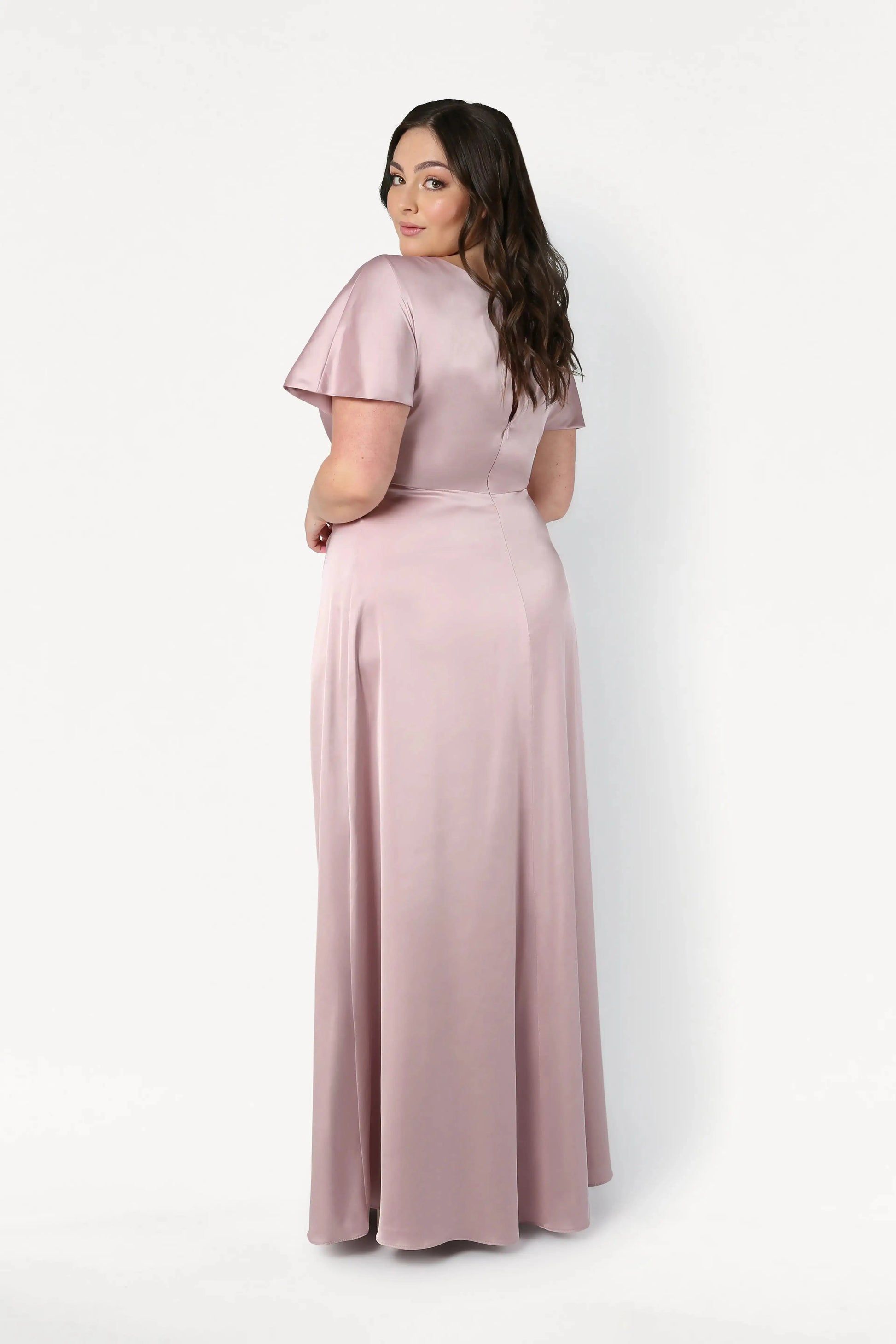 A woman with long brown hair stands slightly turned away, wearing the TH&TH Camilla Bridesmaid Dress in Smoked Blush Satin, a floor-length short-sleeve gown, set against a plain white background.