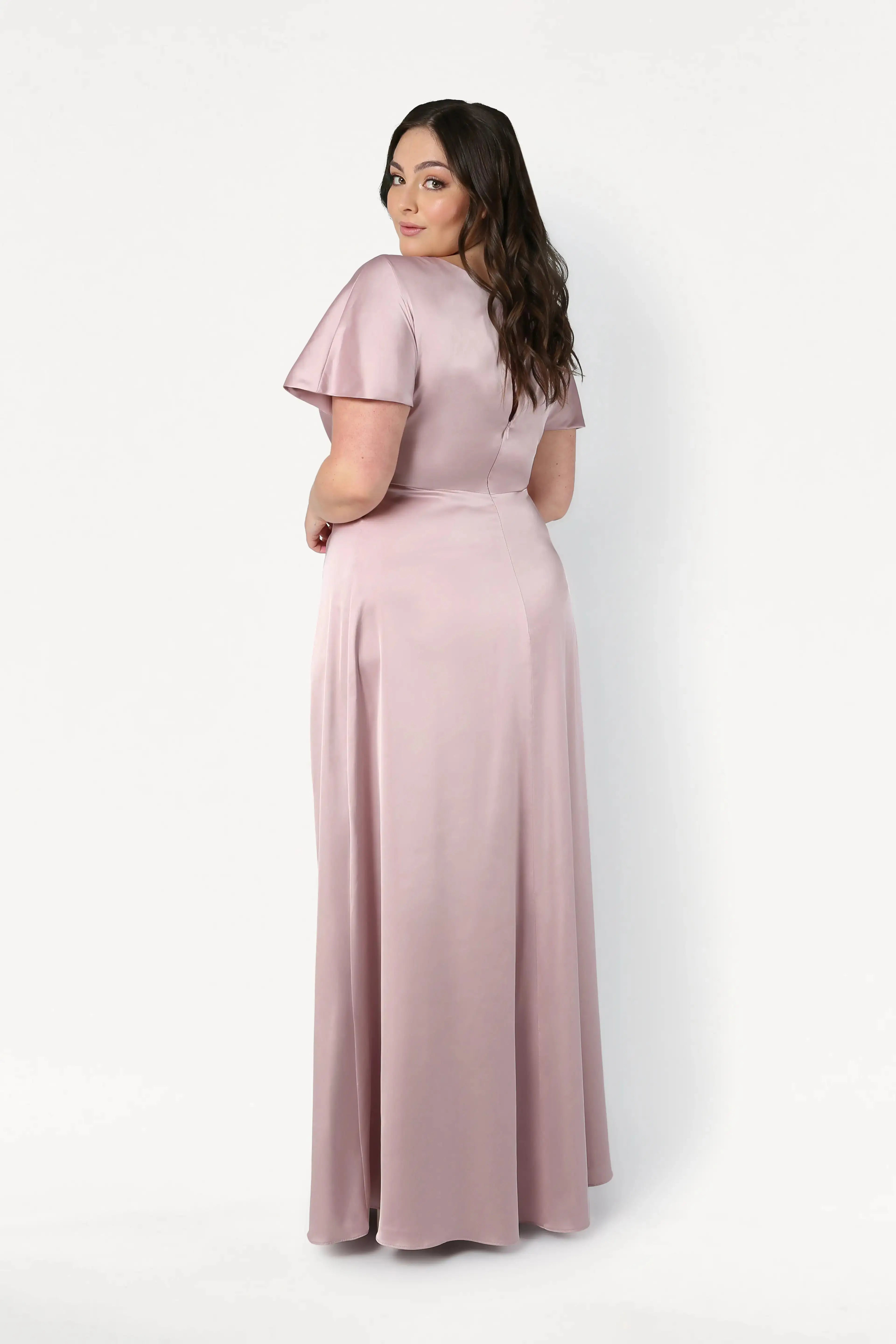 A woman with long brown hair stands slightly turned away, wearing the TH&TH Camilla Bridesmaid Dress in Smoked Blush Satin, a floor-length short-sleeve gown, set against a plain white background.