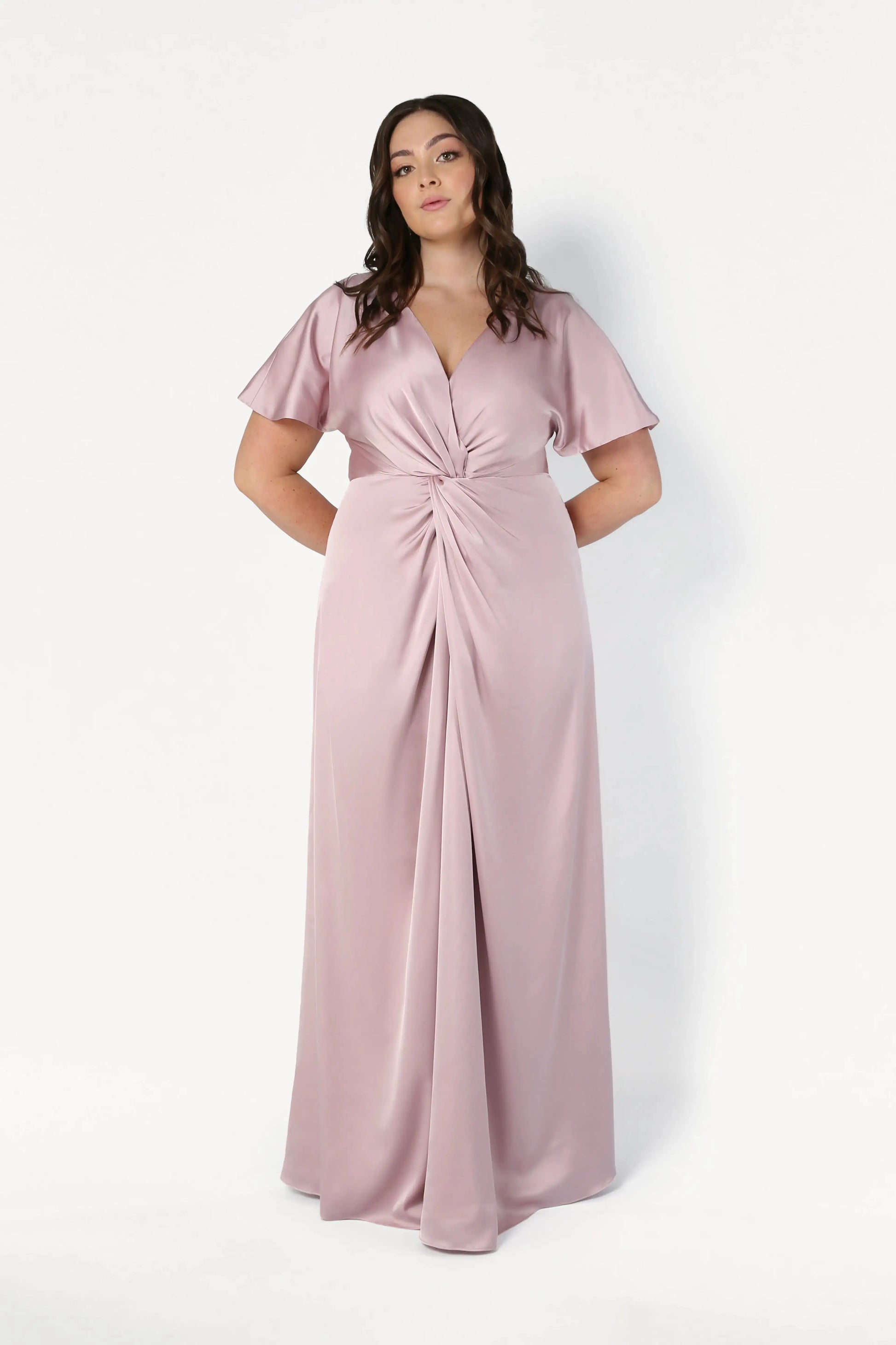 A woman stands against a white background wearing the Camilla Bridesmaid Dress in Smoked Blush Satin by TH&TH, a floor-length gown with short flutter sleeves, V-neckline, and gathered twist waist detail. Her hands are behind her back.