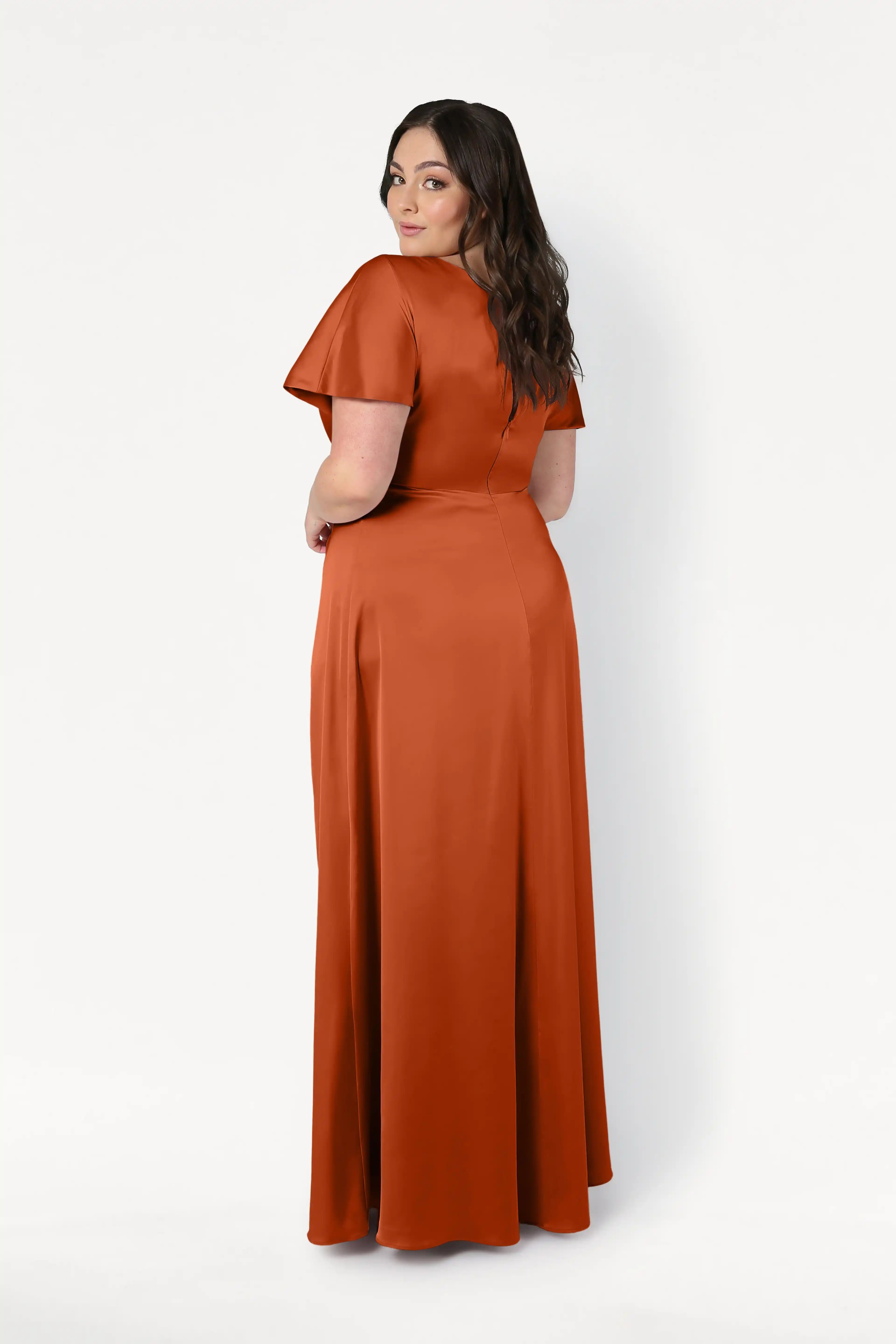 A woman with long, wavy dark hair faces away, looking over her shoulder. She wears the size-inclusive TH&TH Camilla Bridesmaid Dress in Terracotta Satin, a floor-length gown with short sleeves, against a plain white background.