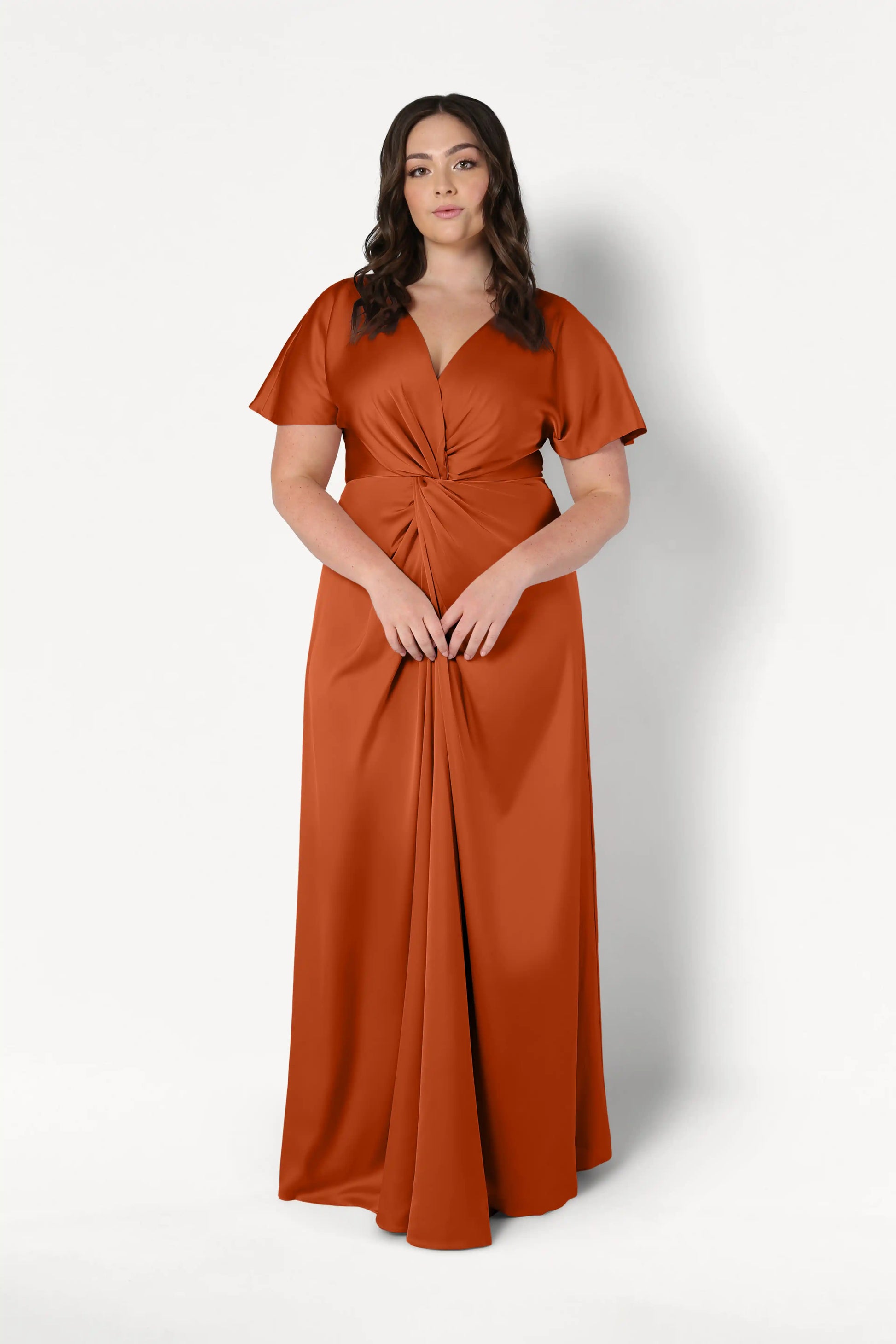 A woman with wavy brown hair wears the TH&TH Camilla Bridesmaid Dress in Terracotta Satin, a floor-length gown with a twist-knot bodice and short sleeves, standing against a plain white background.