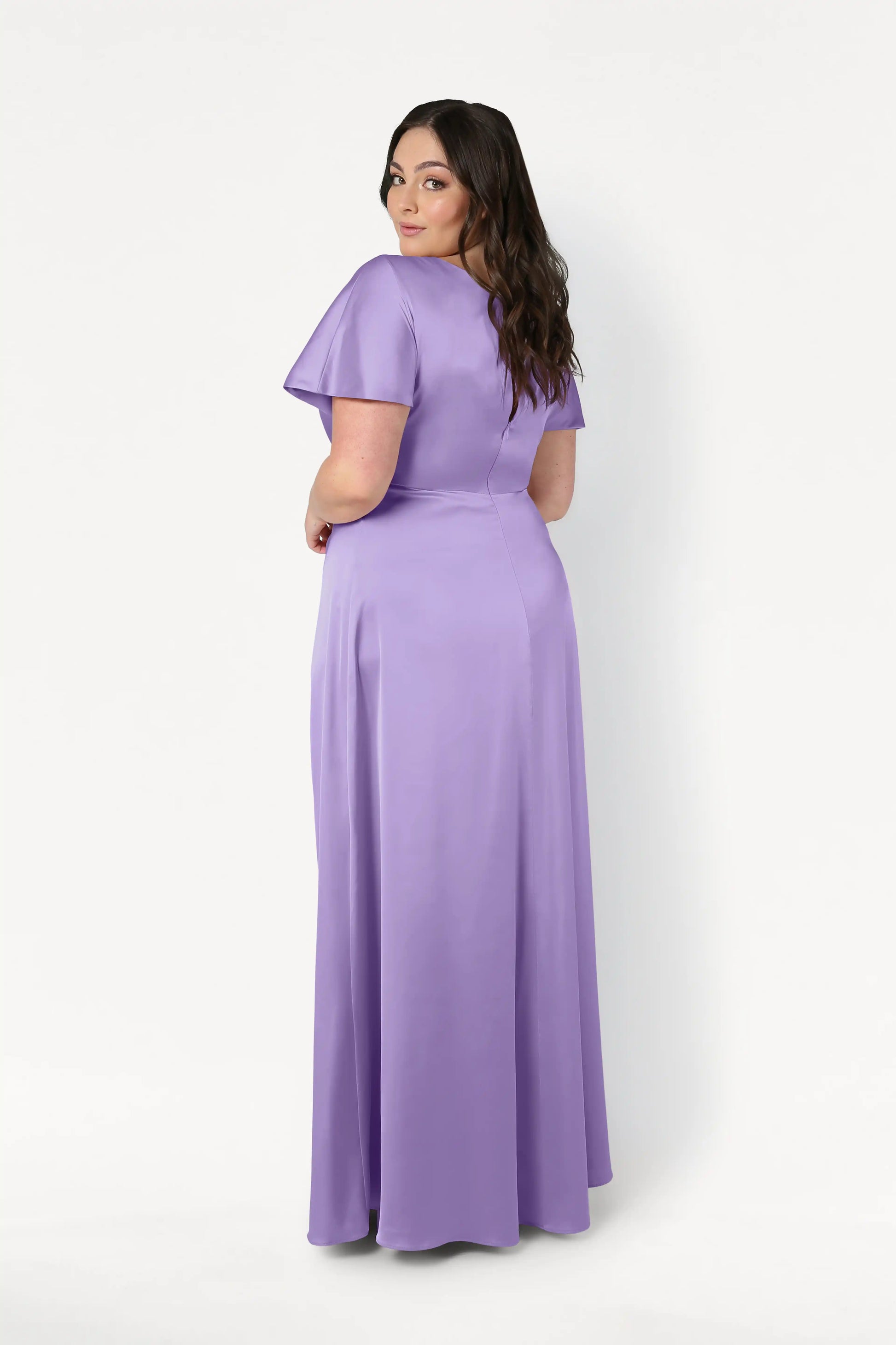A woman with long dark hair poses in the Camilla Bridesmaid Dress in Violet Satin by TH&TH, a size-inclusive floor-length gown with short sleeves, standing against a plain white background and looking over her shoulder at the camera.