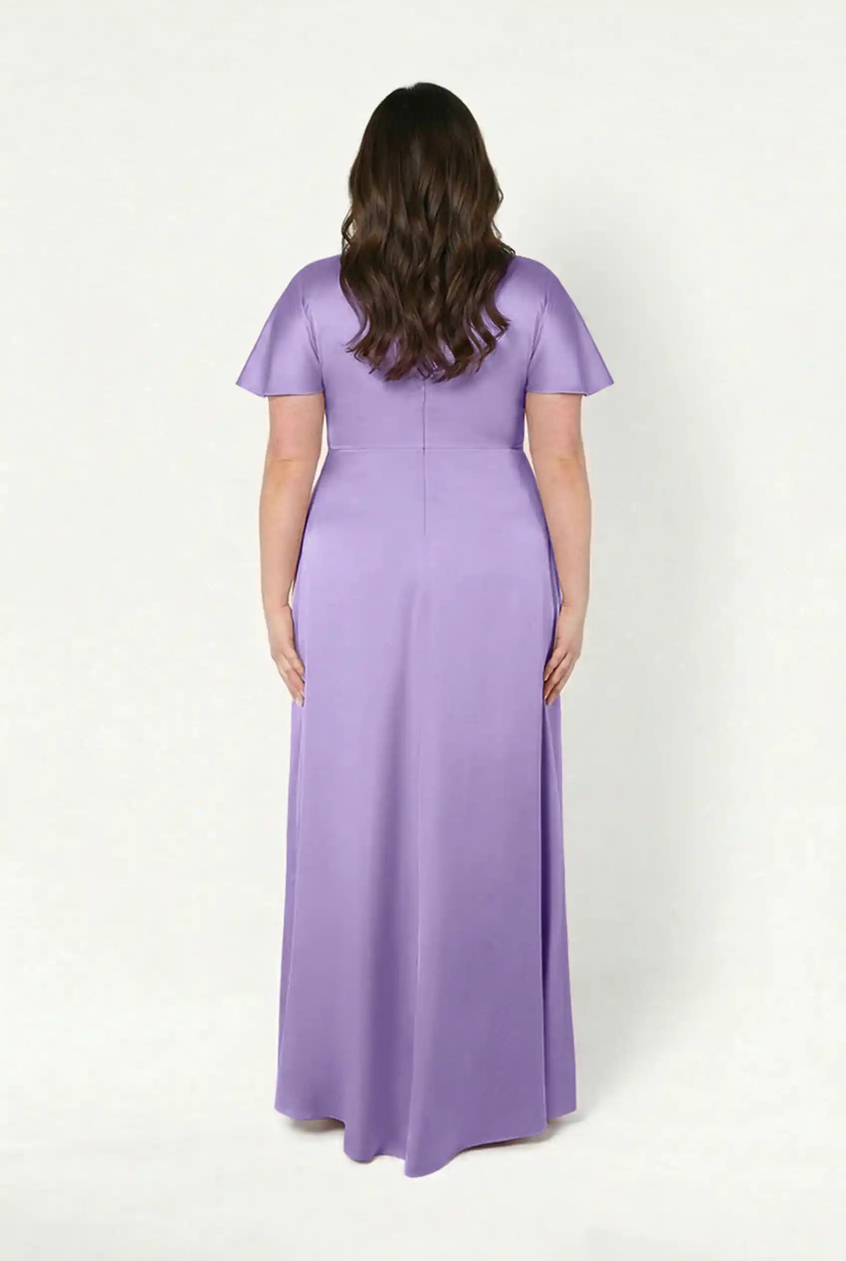 A woman with long brown hair faces away, wearing the TH&TH Camilla Bridesmaid Dress in Violet Satin—a size-inclusive, floor-length gown with short sleeves and a twist-knot bodice—against a plain white background.