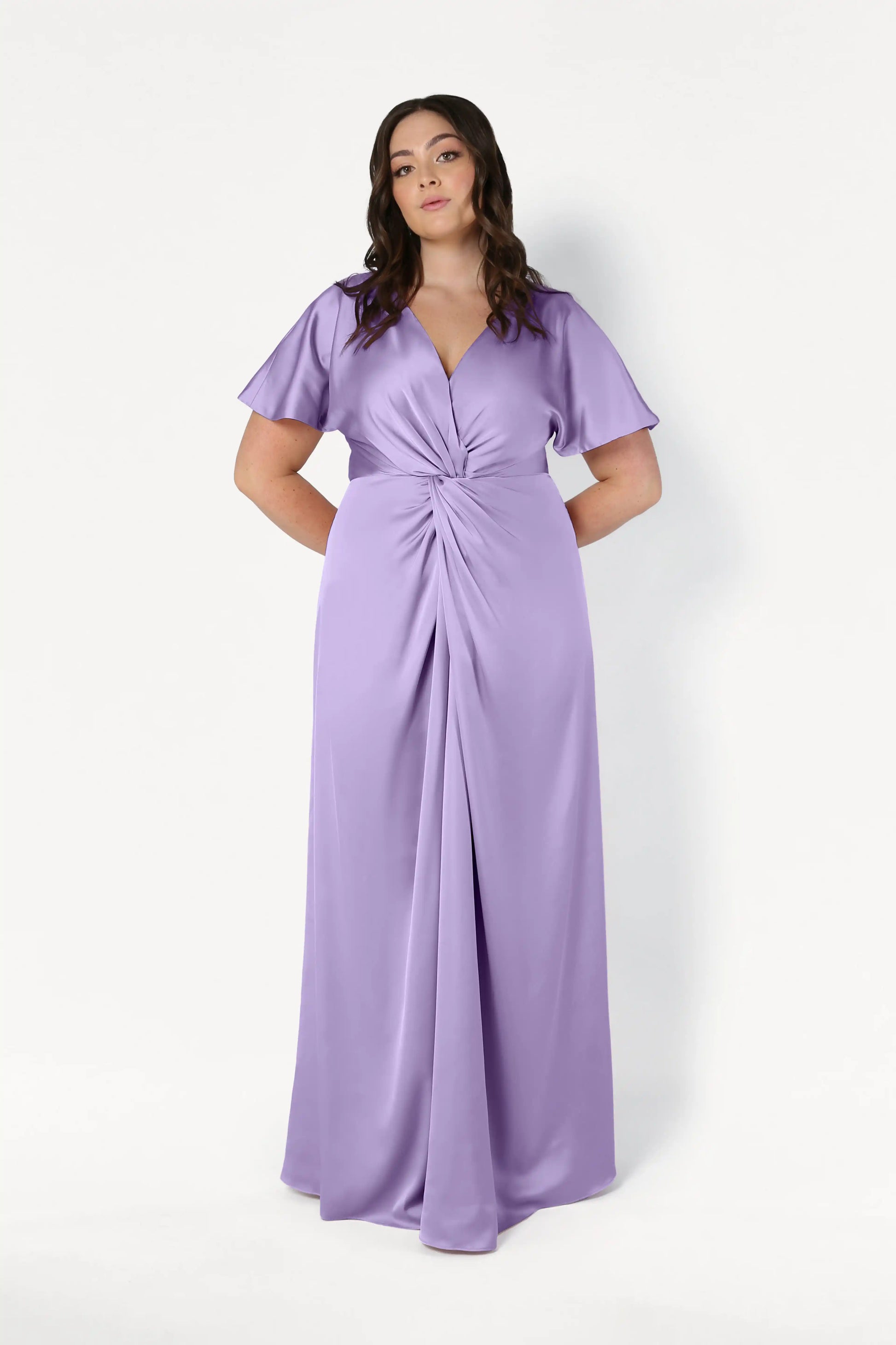 A woman wears the TH&TH Camilla Bridesmaid Dress in Violet Satin, a floor-length gown with short sleeves, V-neckline, and twist-knot bodice, standing against a plain white background.