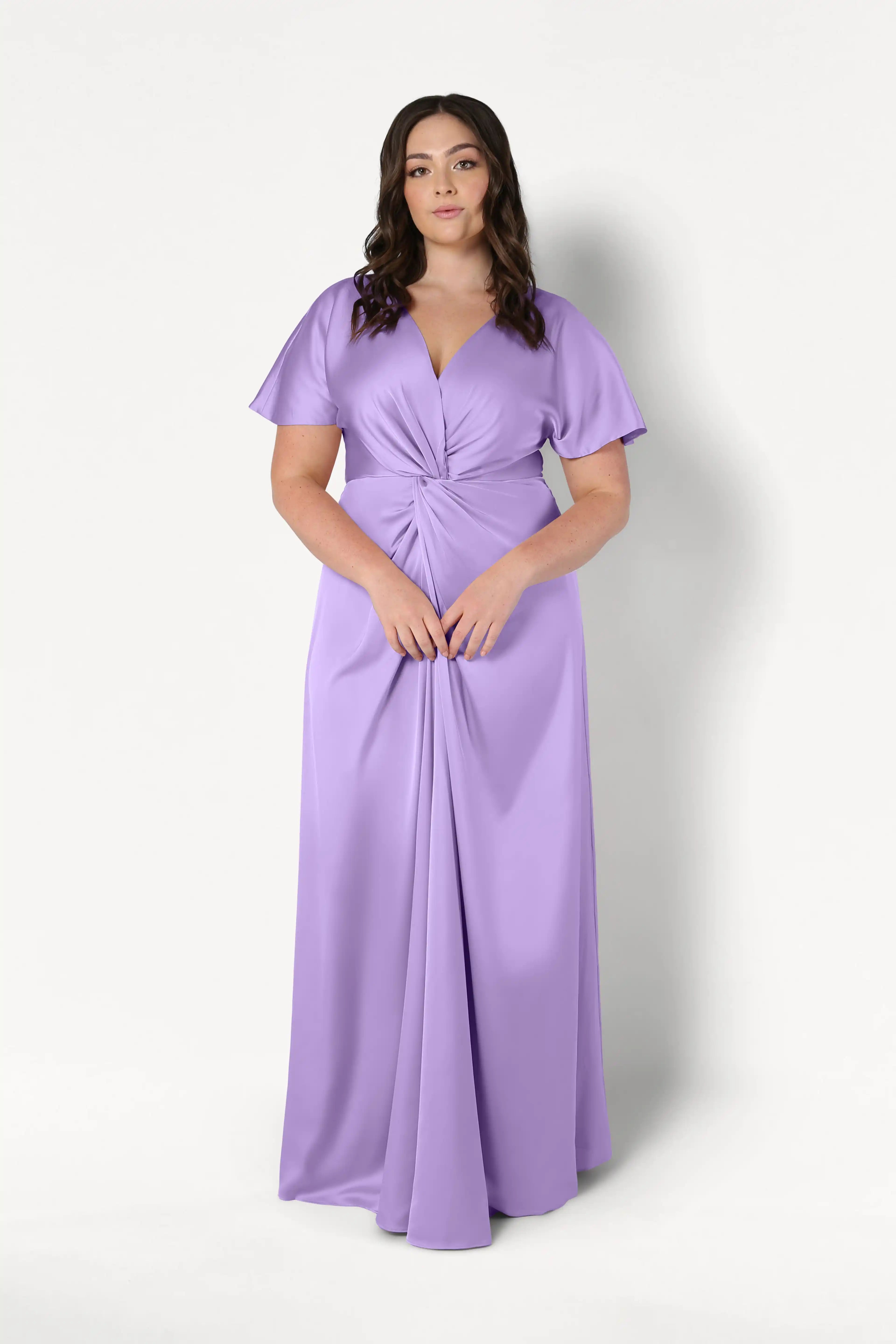 A woman with long brown hair wears the TH&TH Camilla Bridesmaid Dress in violet satin, featuring short sleeves and a twist-knot bodice, standing against a plain white background.