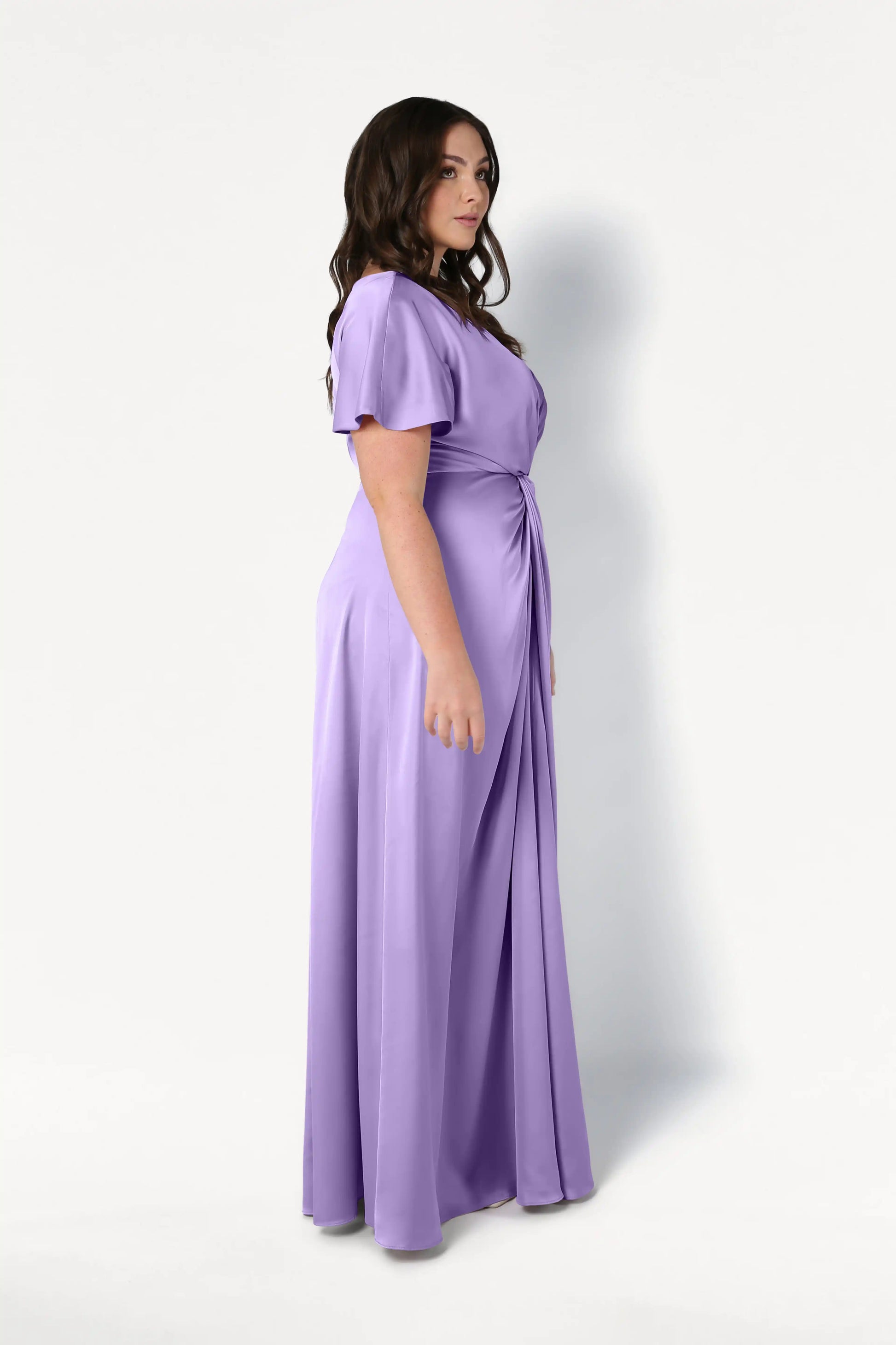 A woman with long dark hair models the TH&TH Camilla Bridesmaid Dress in violet satin, featuring short sleeves and a twist-knot bodice, standing sideways against a plain white background.