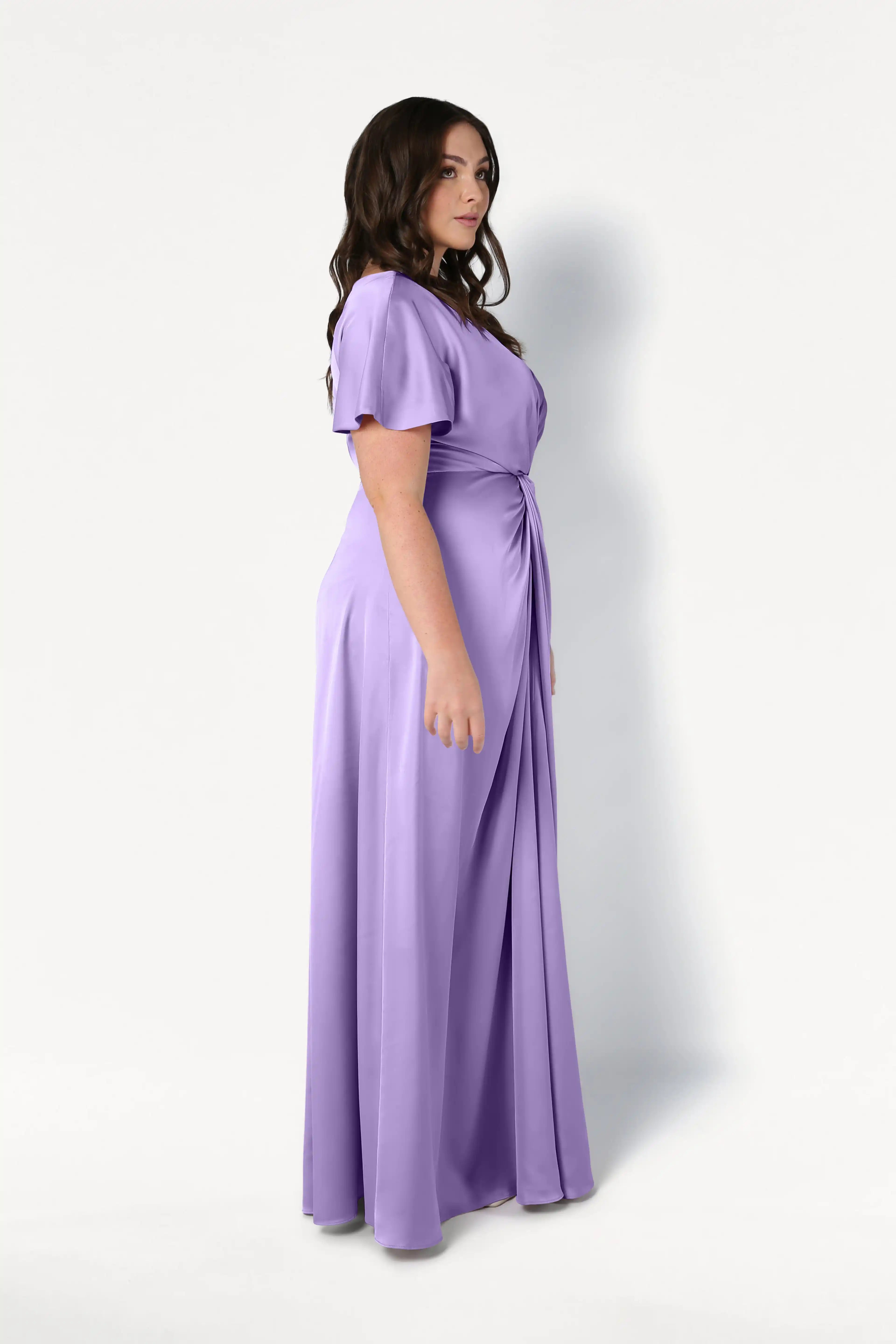 A woman with long dark hair models the TH&TH Camilla Bridesmaid Dress in violet satin, featuring short sleeves and a twist-knot bodice, standing sideways against a plain white background.