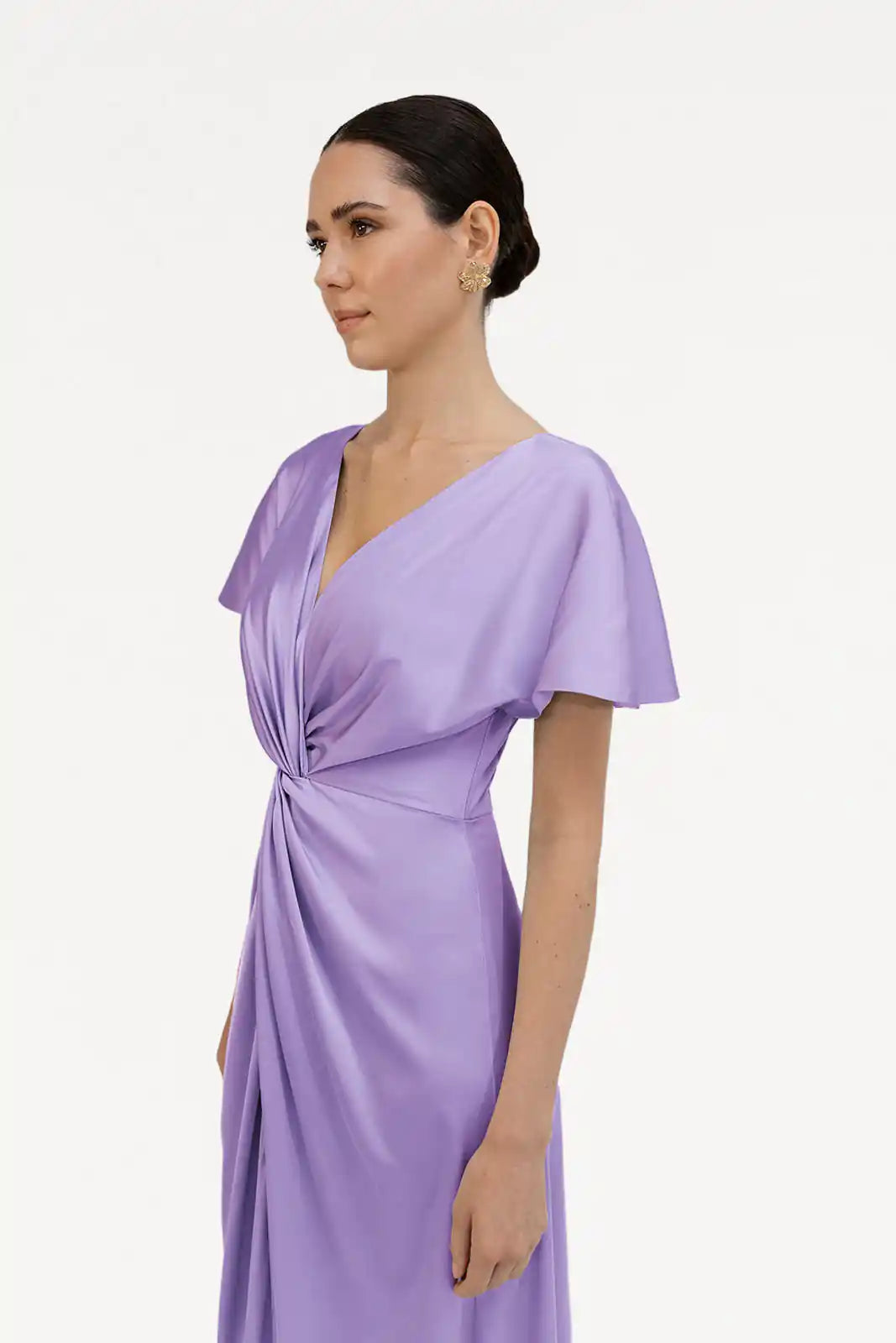 A woman with dark hair in a low bun, wearing gold earrings and the size-inclusive TH&TH Camilla Bridesmaid Dress in Violet Satin with flutter sleeves, stands against a plain white background, gazing slightly to the side.