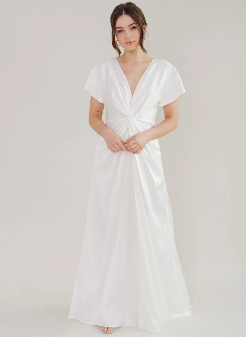 A woman with loosely styled brown hair and a calm expression wears the TH & TH Camilla Dress in Ivory Satin, a floor-length bridesmaid gown featuring short sleeves, a deep V-neckline, and twist-knot detail at the waist against a plain light background.