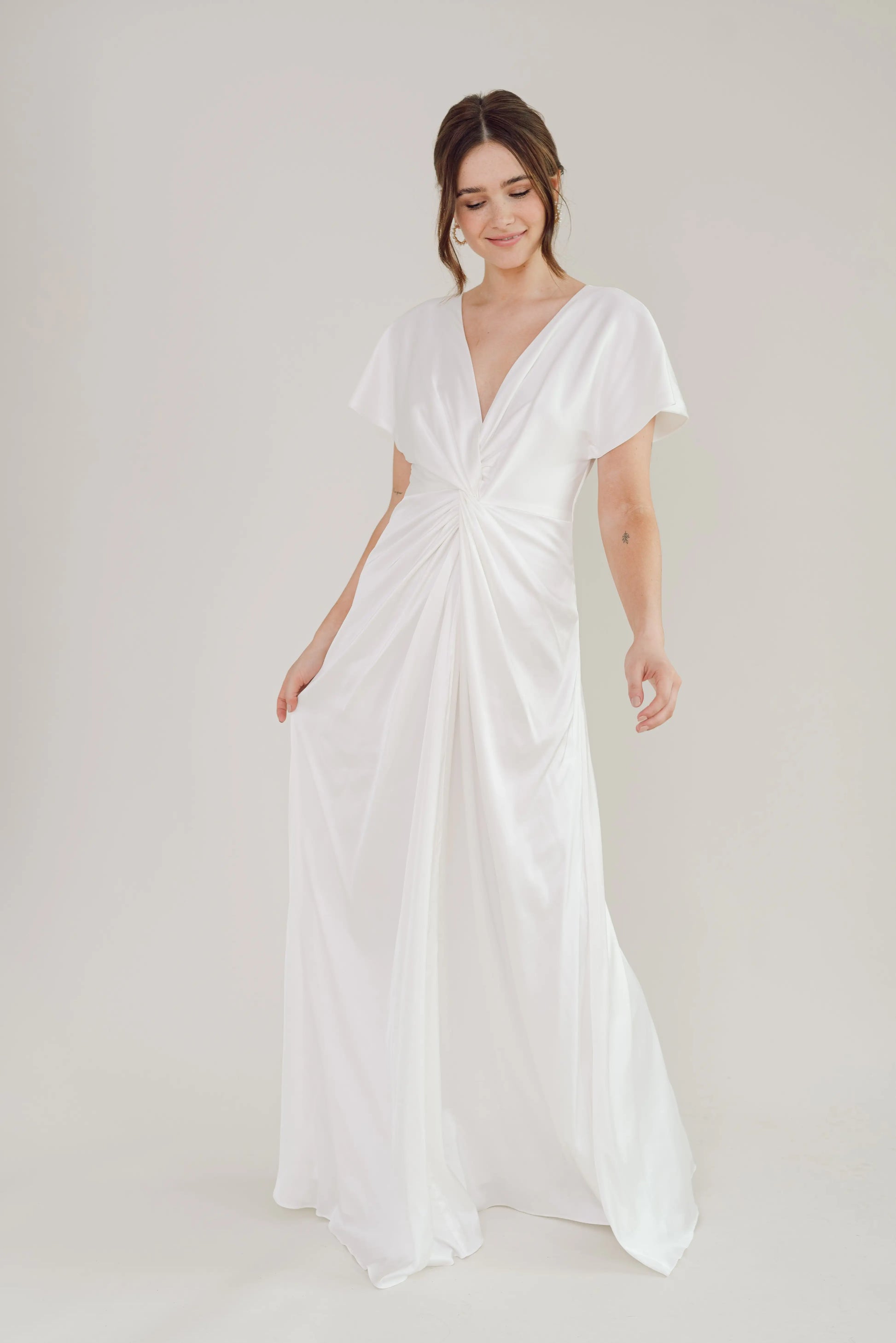 A woman wears the Camilla Dress in Ivory Satin by TH & TH, a size-inclusive gown with short sleeves and a deep V-neckline, standing against a plain background and softly smiling as she gently holds her dress.