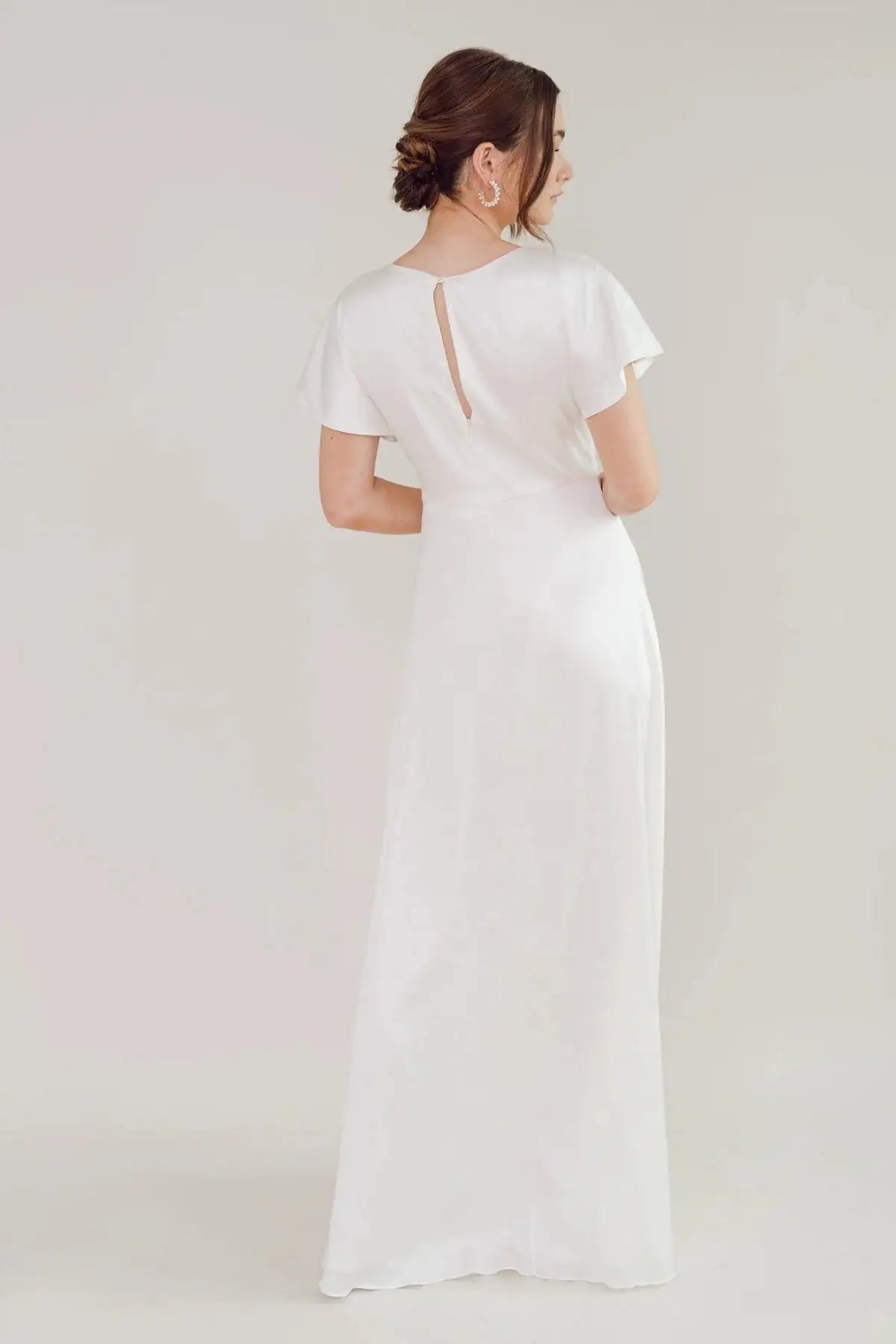 A woman with brown hair in an updo is seen from the back, wearing the TH & TH Camilla Dress in Ivory Satin—a floor-length bridesmaid gown with short sleeves and a twist-knot bodice—against a plain light background.