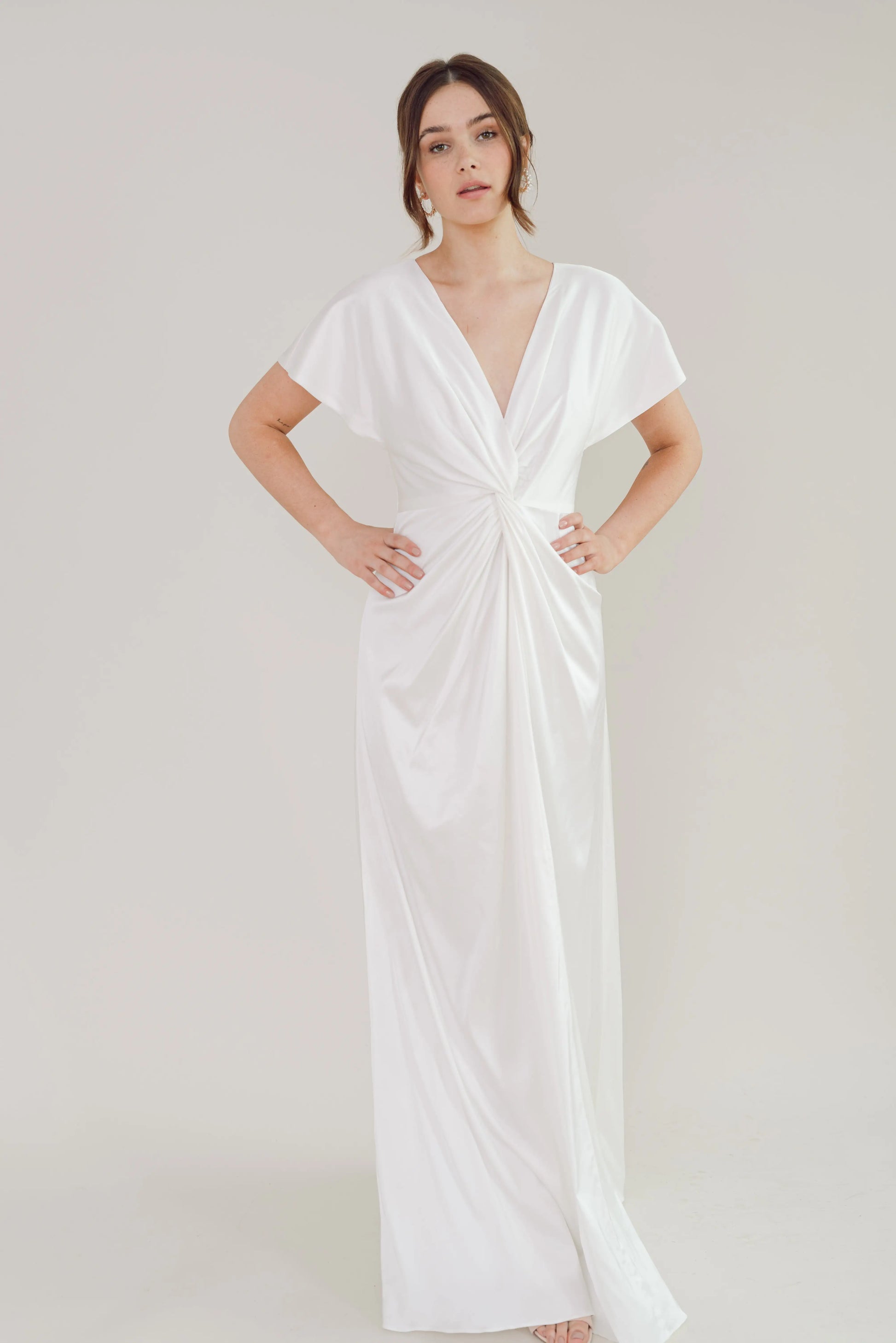 A woman stands against a plain light background, wearing the Camilla Dress in Ivory Satin by TH & TH—a size-inclusive bridesmaid gown featuring short sleeves, a deep V-neck, and a twist-knot bodice. Her hands rest on her hips as she looks forward.