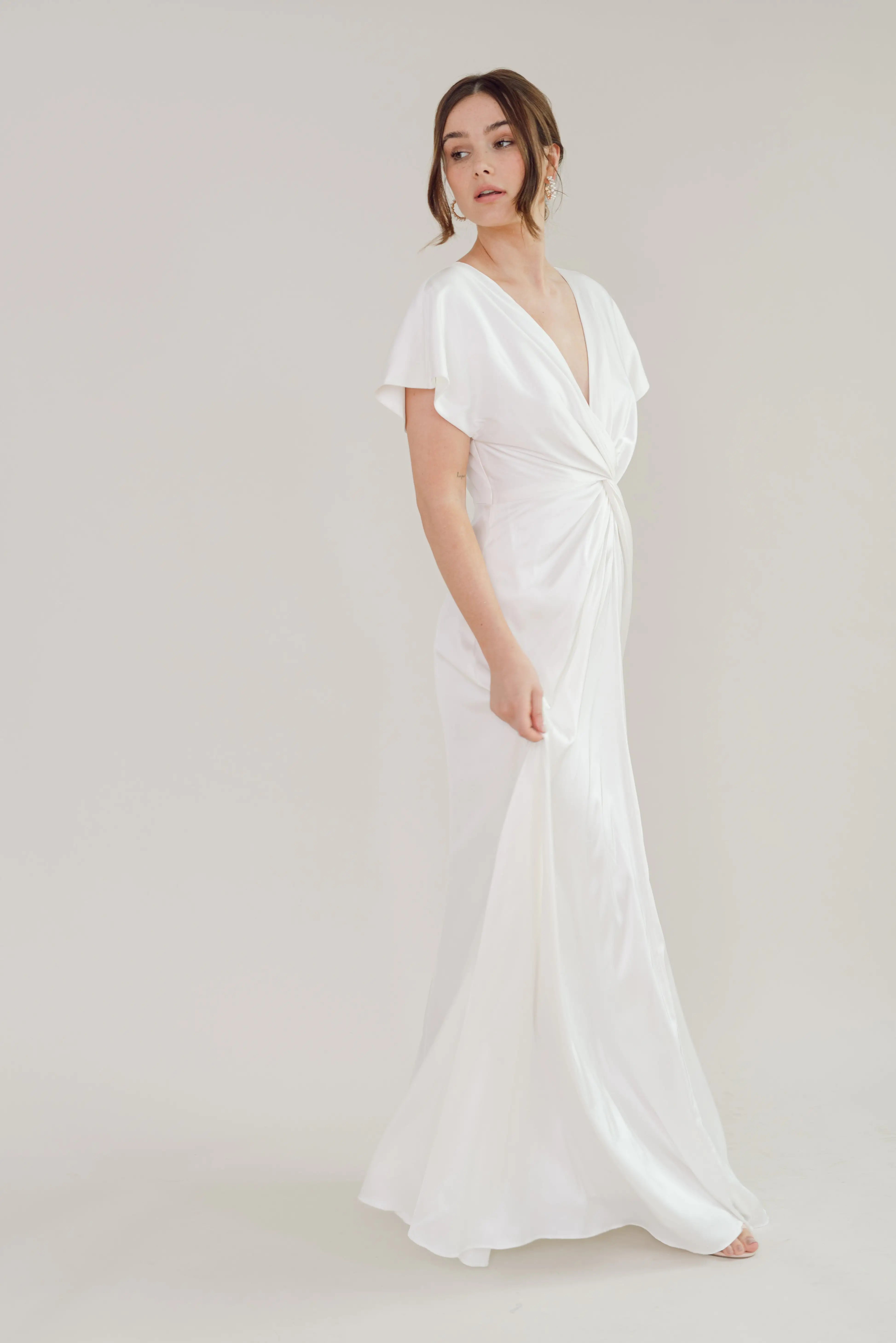 A woman in the Camilla Dress in Ivory Satin by TH & TH—featuring a deep V-neckline, short sleeves, and size-inclusive design—stands against a plain light background, gently holding the elegant gown as she gazes thoughtfully aside.