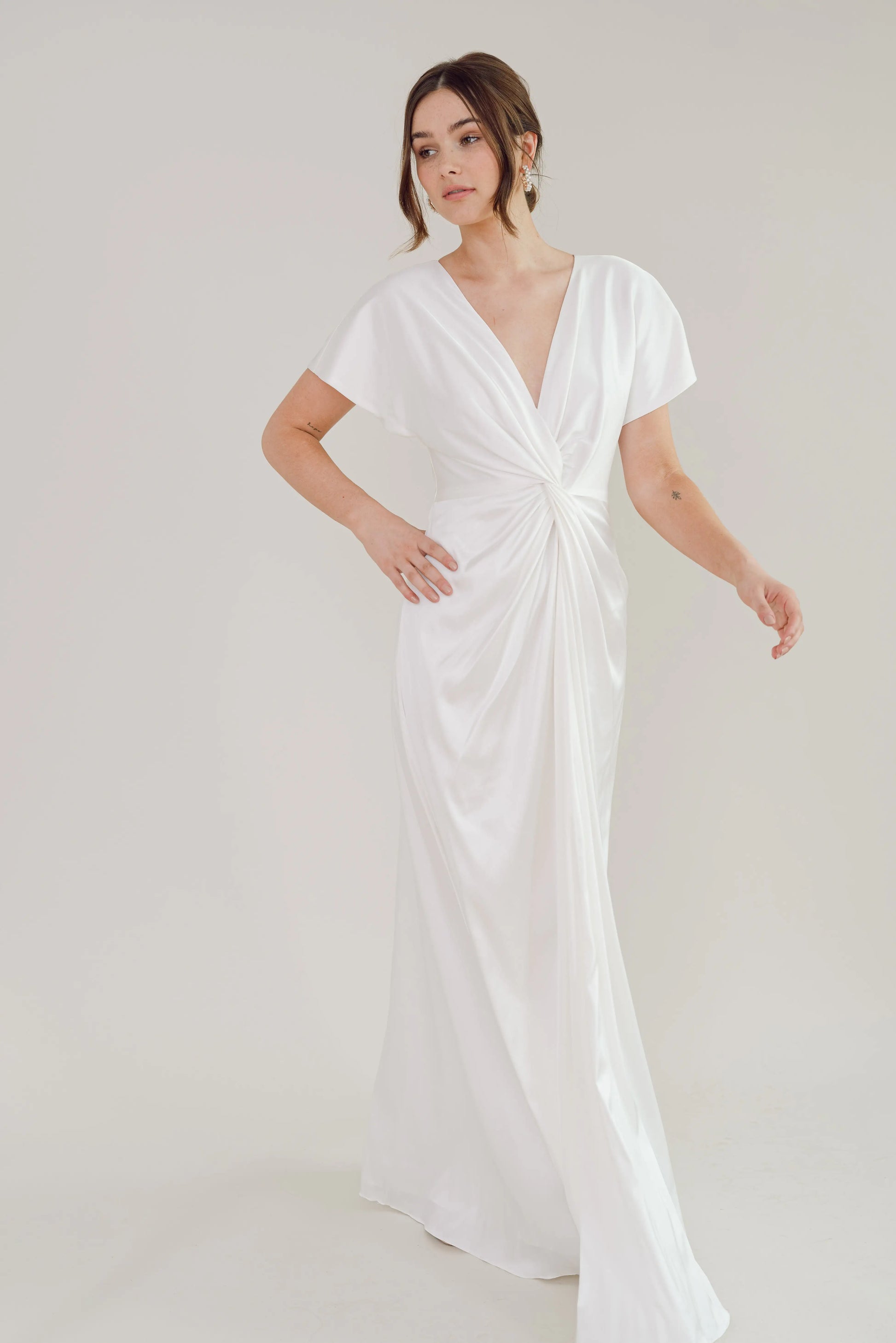 A woman models the TH & TH Camilla Dress in Ivory Satin, a size-inclusive, floor-length bridesmaid gown with short sleeves, a deep V-neckline, and a twist-knot bodice that gathers at the waist; shown against a light backdrop.