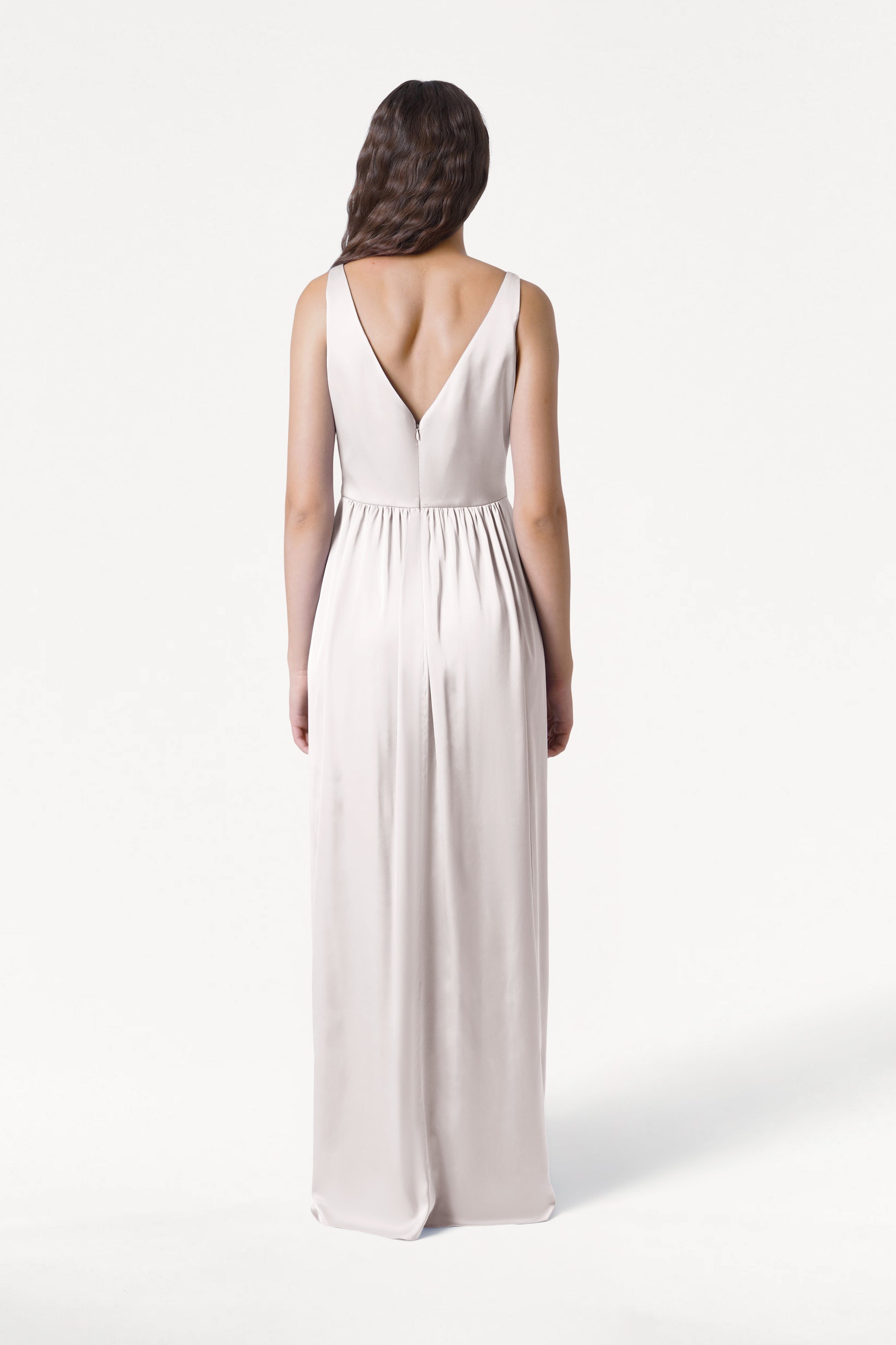 A woman with long, wavy brown hair stands facing away in the sleeveless Carmen Bridesmaid Dress in Oyster Satin by TH&TH, featuring a V-shaped back—perfect for modern bridal parties—set against a plain white background.