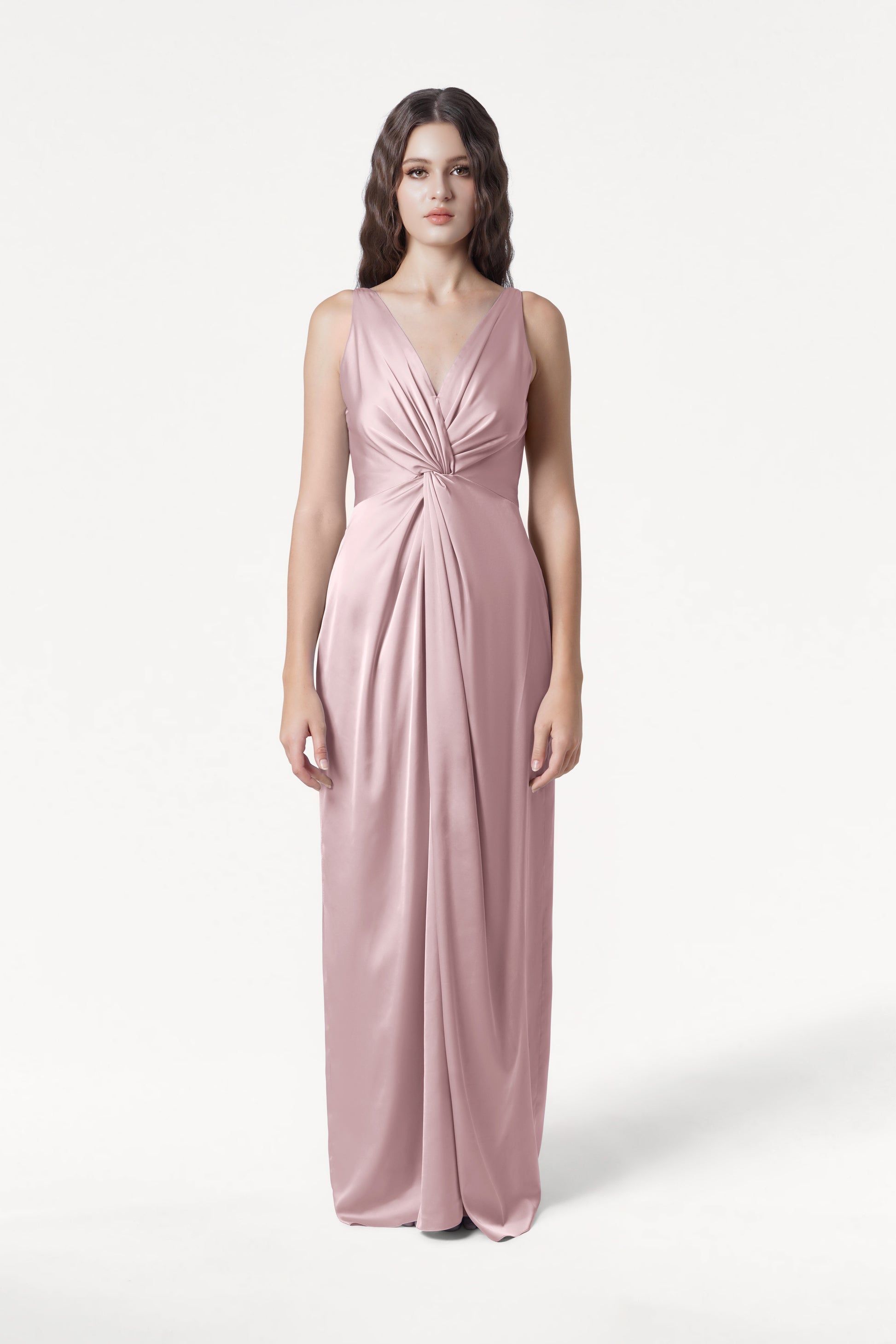 A woman poses against a white background in the TH&TH Carmen Bridesmaid Dress—an elegant, sleeveless, floor-length smoked blush satin gown with a deep V-neckline and gathered waist, ideal for modern bridal parties.
