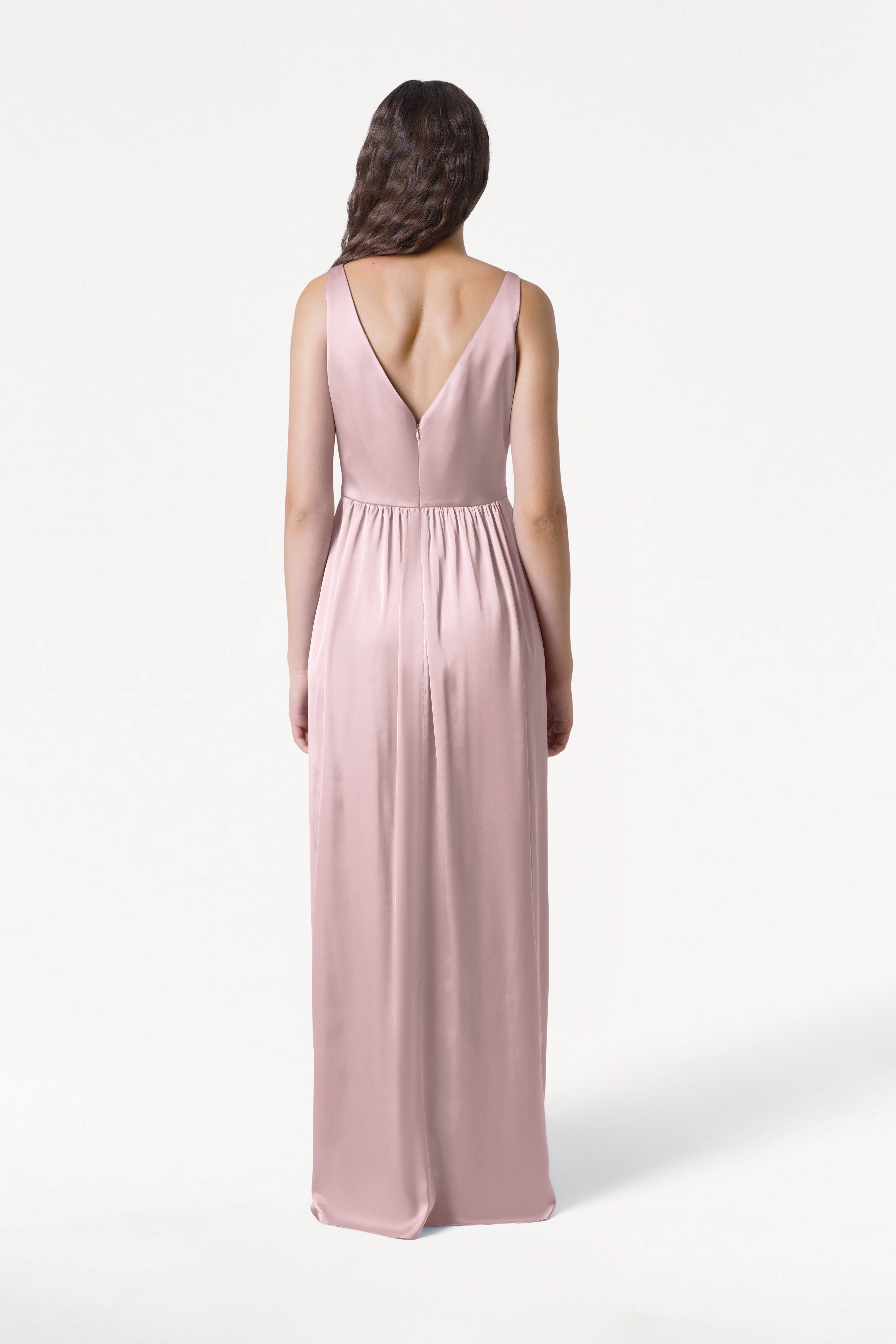 A woman faces away, modeling the TH&TH Carmen Bridesmaid Dress in Smoked Blush Satin—a long, sleeveless gown with a deep V-back and gathered waist. Her loose waves complete this elegant look, ideal for modern bridal parties.