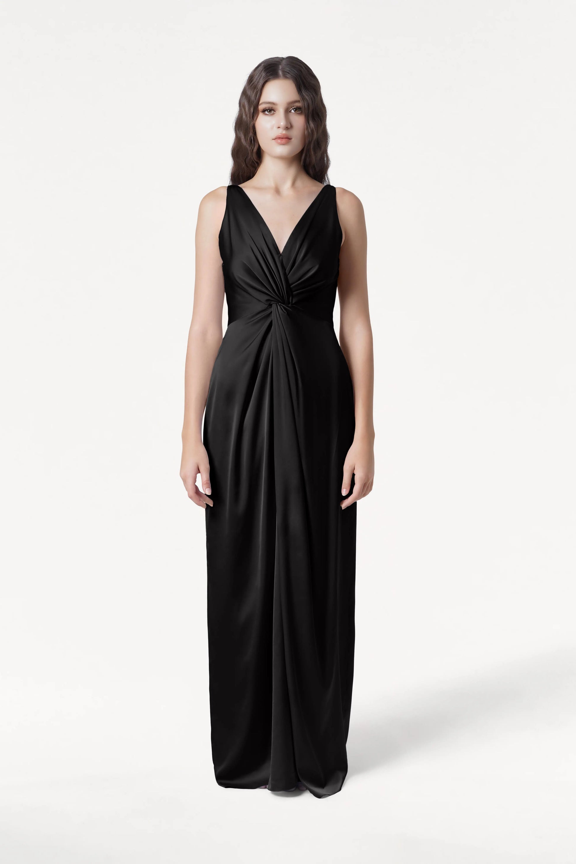 A woman faces forward in the TH&TH Carmen Bridesmaid Dress in Black Satin—a sleeveless, floor-length gown with a deep V-neck and gathered waist—her long wavy brown hair and neutral expression set against a plain white background.