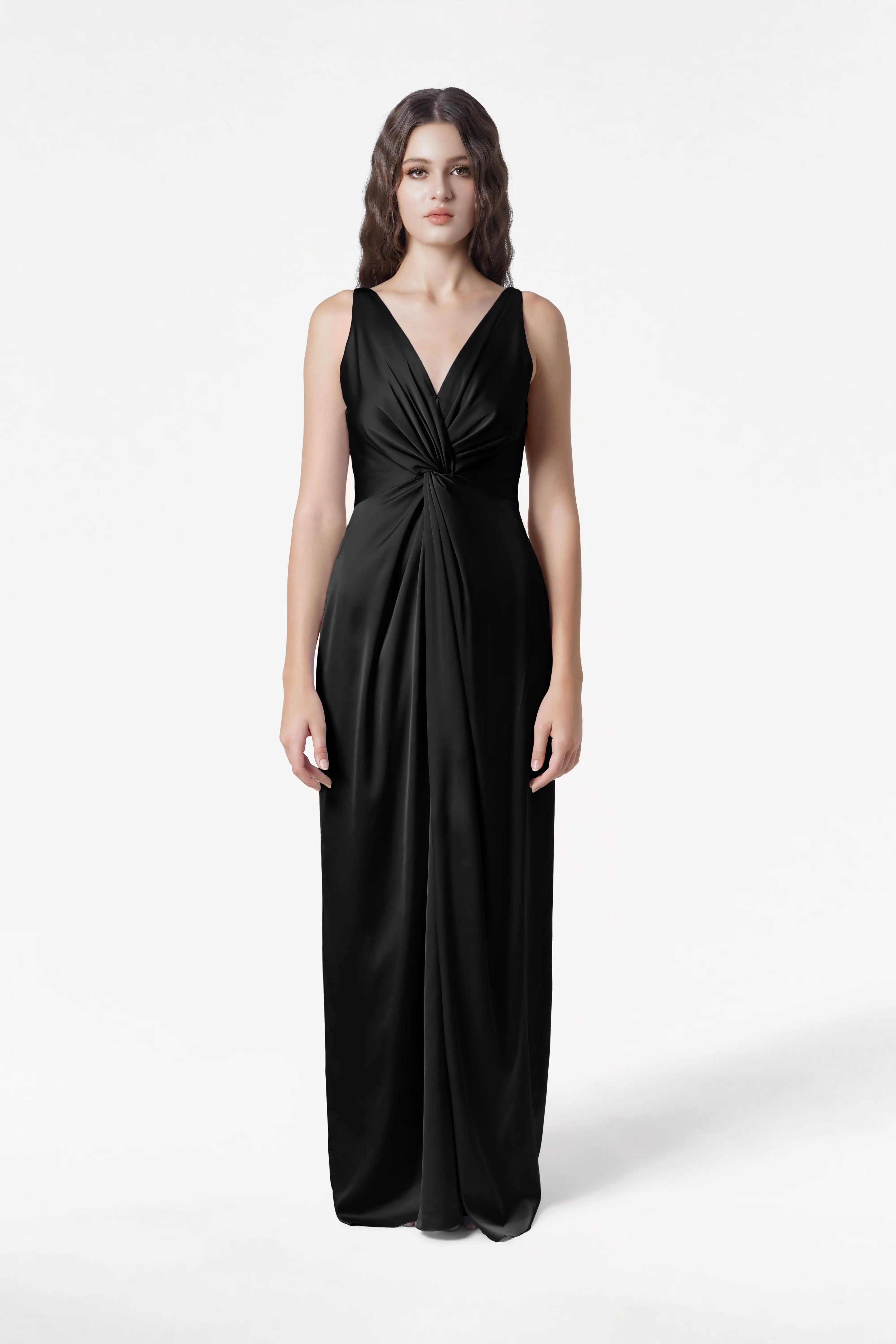 A woman faces forward in the TH&TH Carmen Bridesmaid Dress in Black Satin—a sleeveless, floor-length gown with a deep V-neck and gathered waist—her long wavy brown hair and neutral expression set against a plain white background.